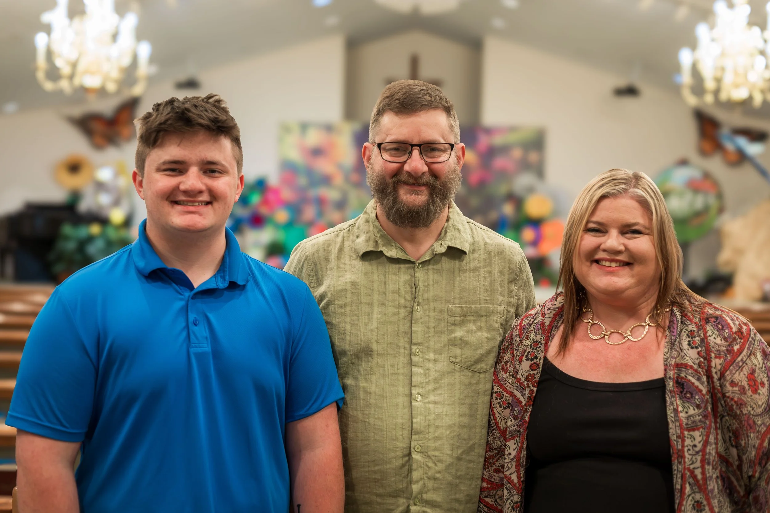 Three people standing inside a church, smiling at the camera. A young man in a blue polo shirt on the left, a bearded man with glasses and a green shirt in the middle, and a woman with blonde hair wearing a patterned jacket and black top on the right. The background shows church decorations and a cross on the wall.