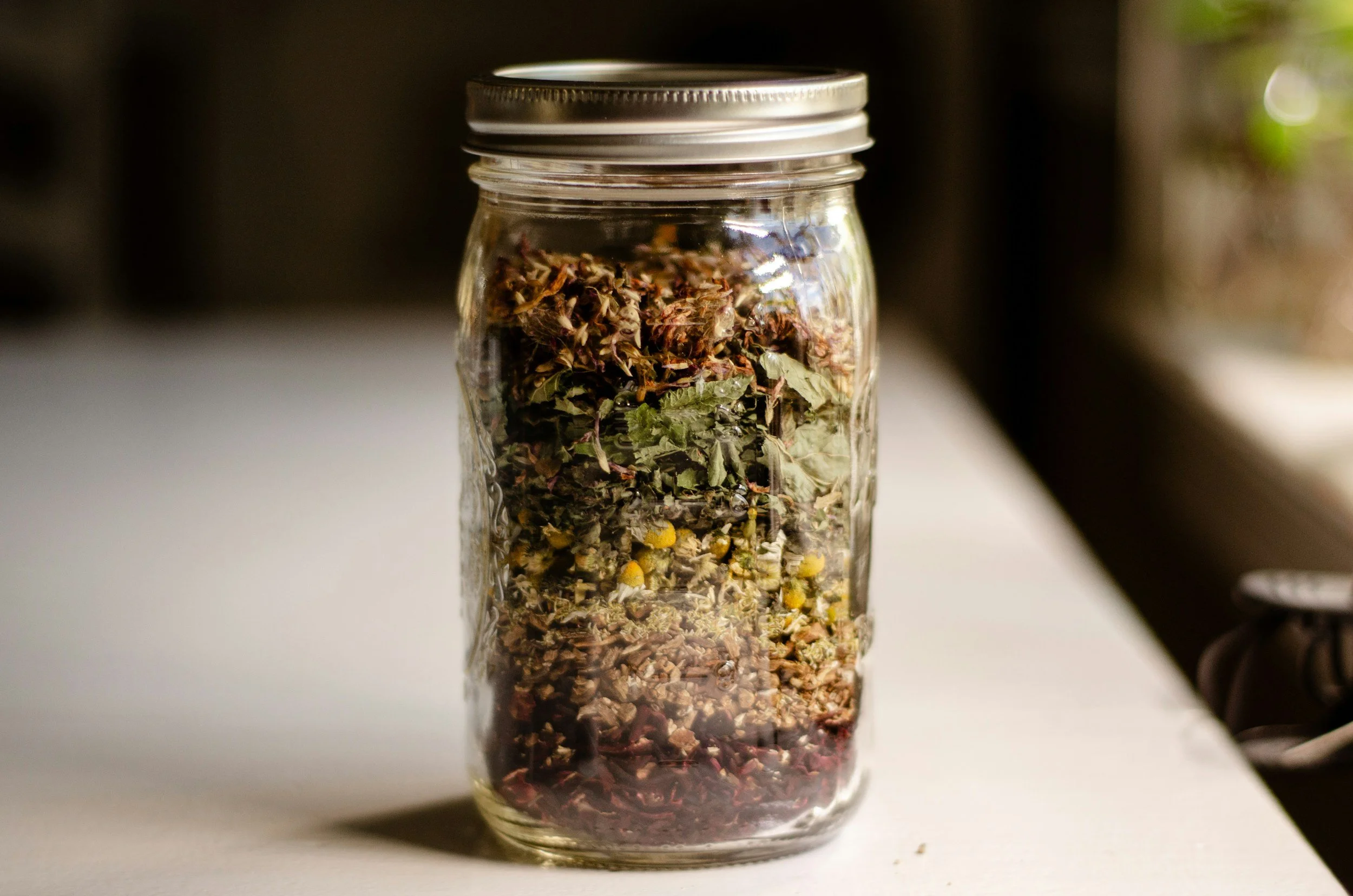 A glass jar filled with dried herbs and flowers on a white surface, with a blurred background.