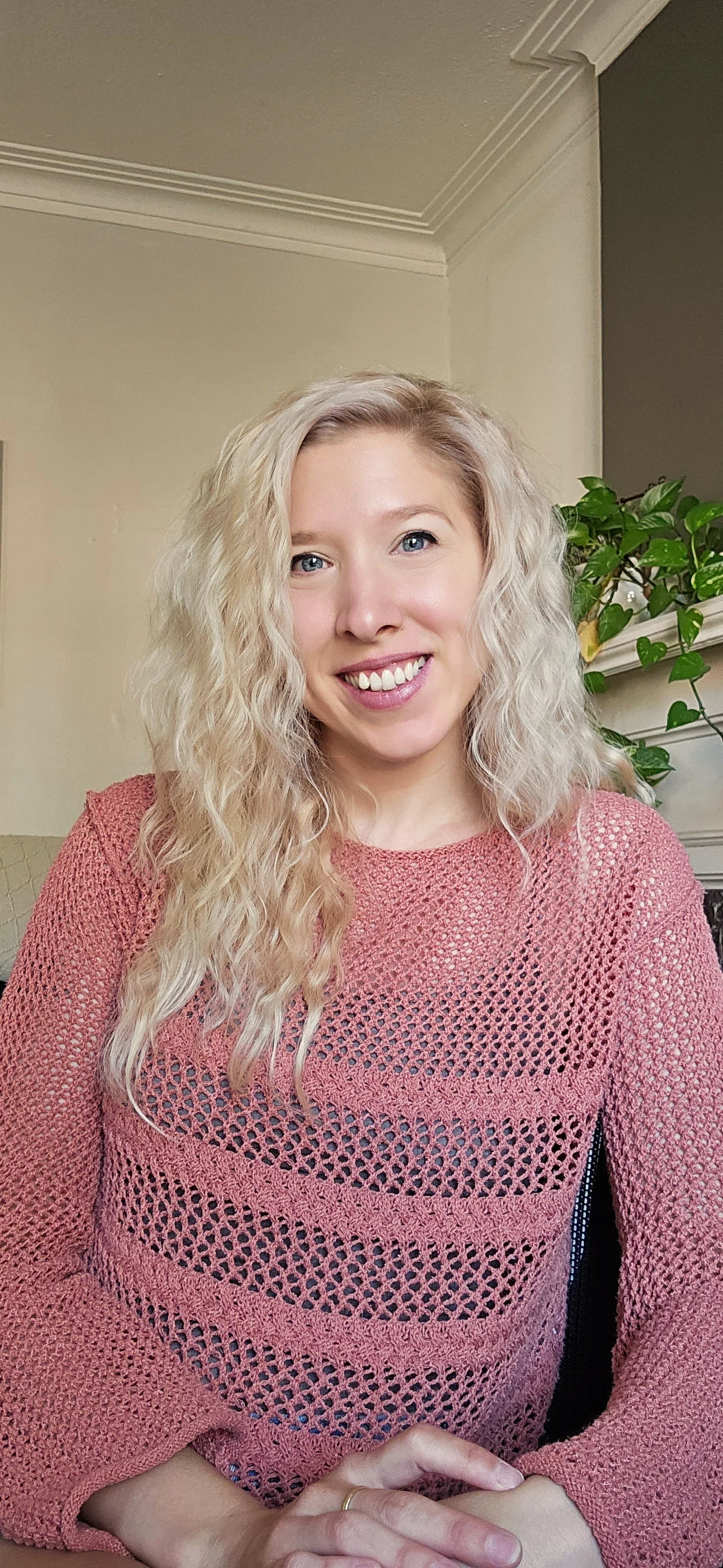 A woman with long, curly blonde hair and blue eyes, smiling, wearing a pink, open-knit sweater, seated indoors with a green plant and neutral-colored walls in the background.