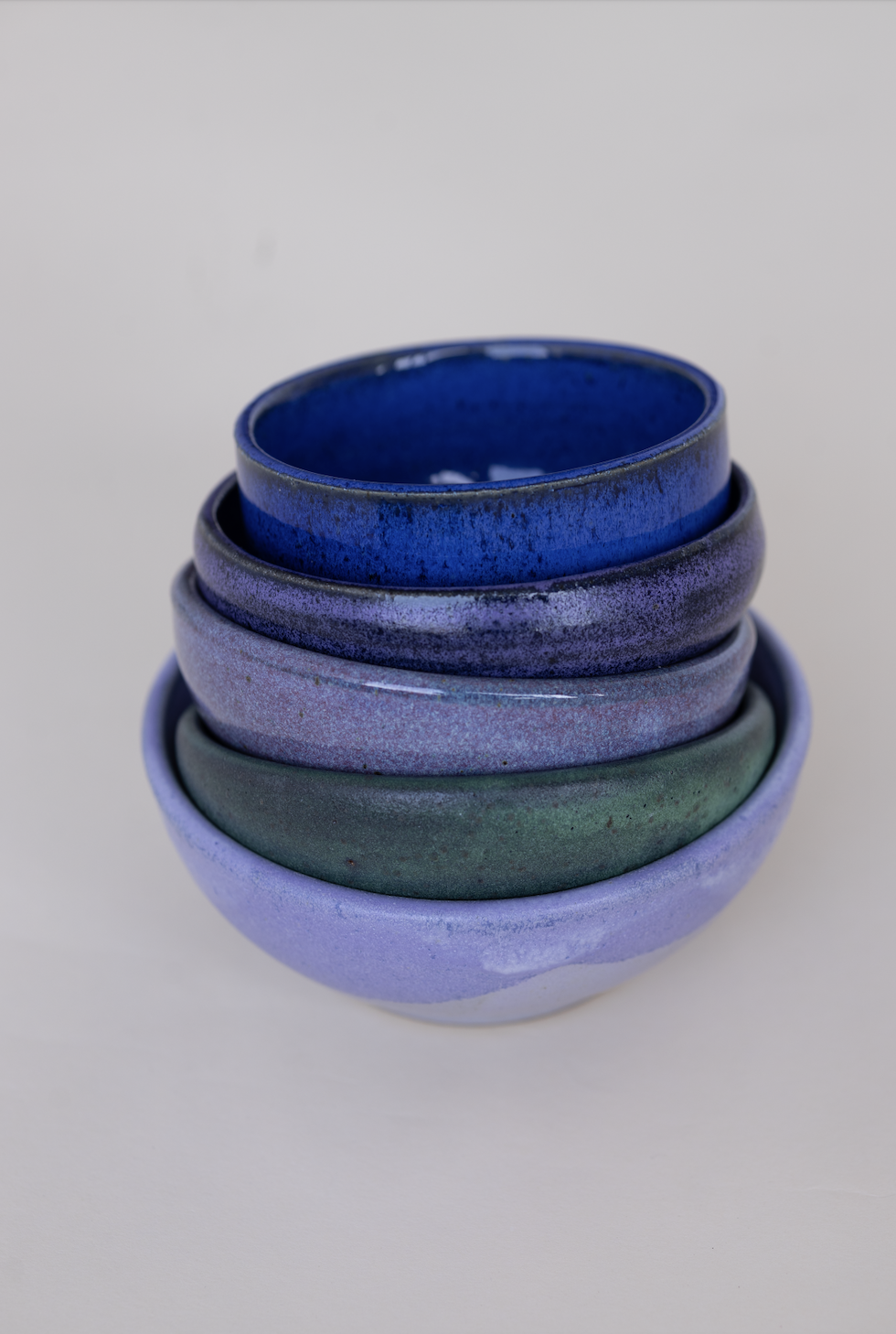 A stack of five colorful ceramic bowls, from top to bottom: dark blue, purple, lavender, green, and light purple, placed on a plain white surface.