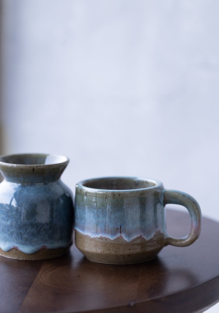Two handmade ceramic mugs with blue and earthy glaze, placed on a wooden surface against a blurred light background.