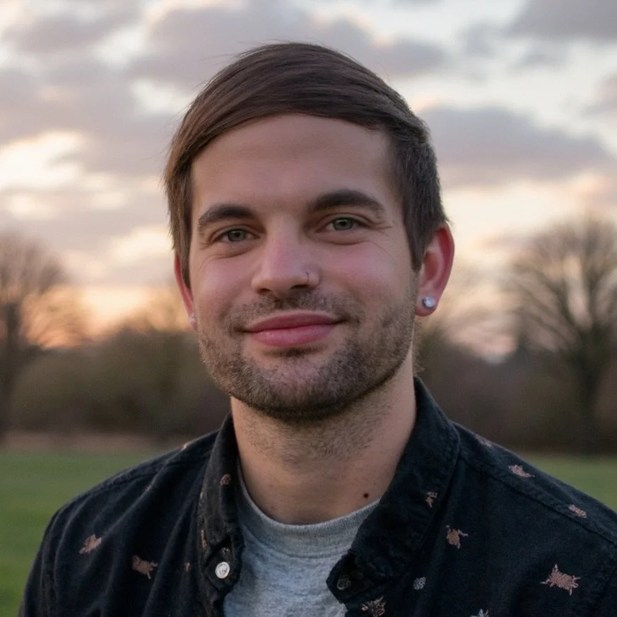 Portrait of a young man with short brown hair, a nose piercing, and earrings, standing outdoors during sunset with trees in the background.
