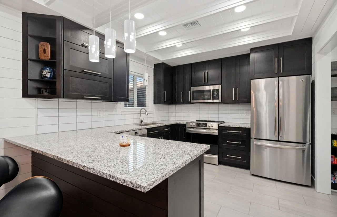 Modern kitchen with black cabinets, stainless steel appliances, granite countertop, white tiled backsplash, and pendant lighting.