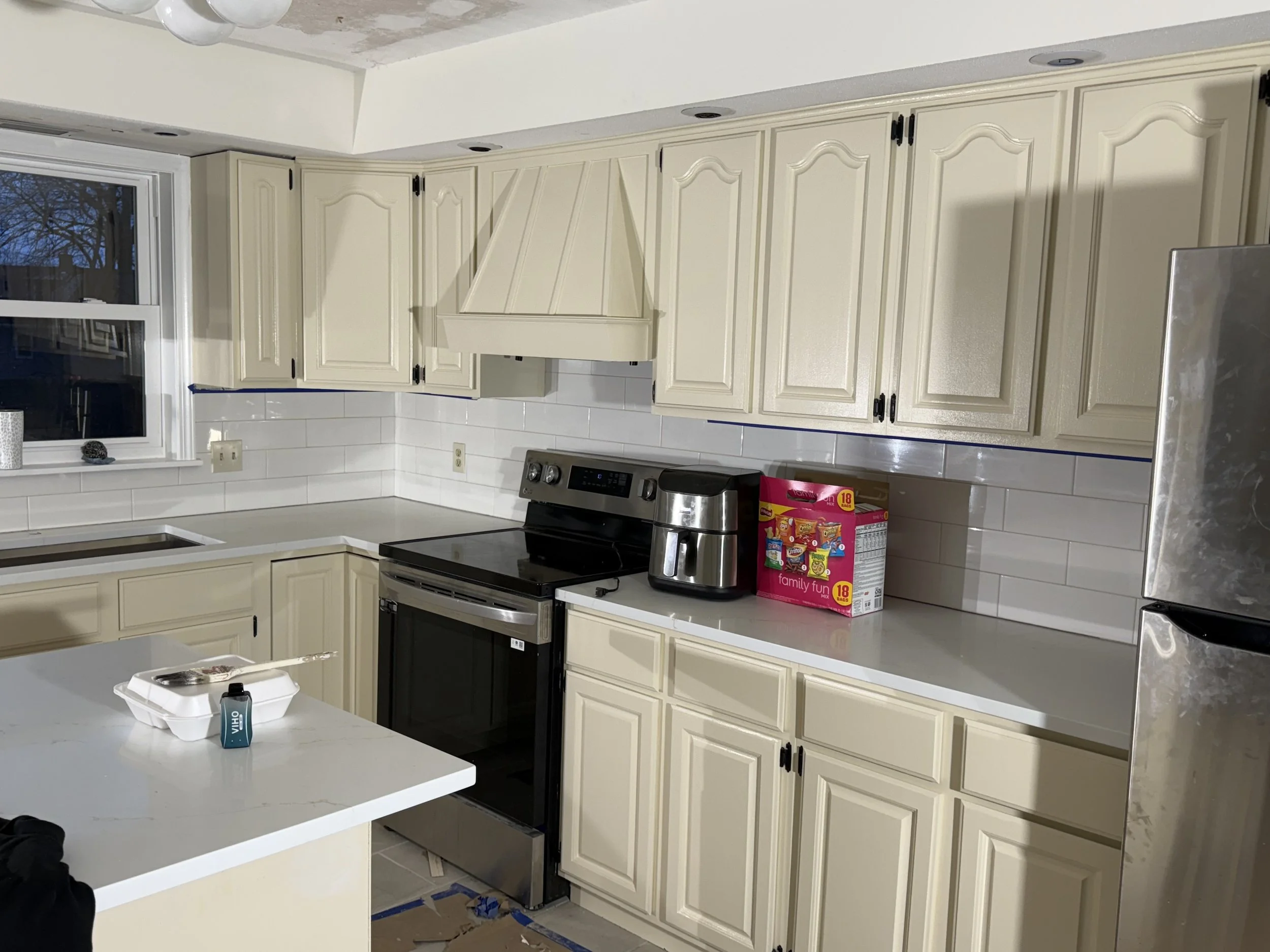 Kitchen with cream-colored cabinets, black stove and oven, stainless steel refrigerator, white countertop, and white tiled backsplash. A window above the sink shows a tree outside. Items on the counter include a pink box, a coffee maker, and a container of food with a spoon and a bottle on the island.