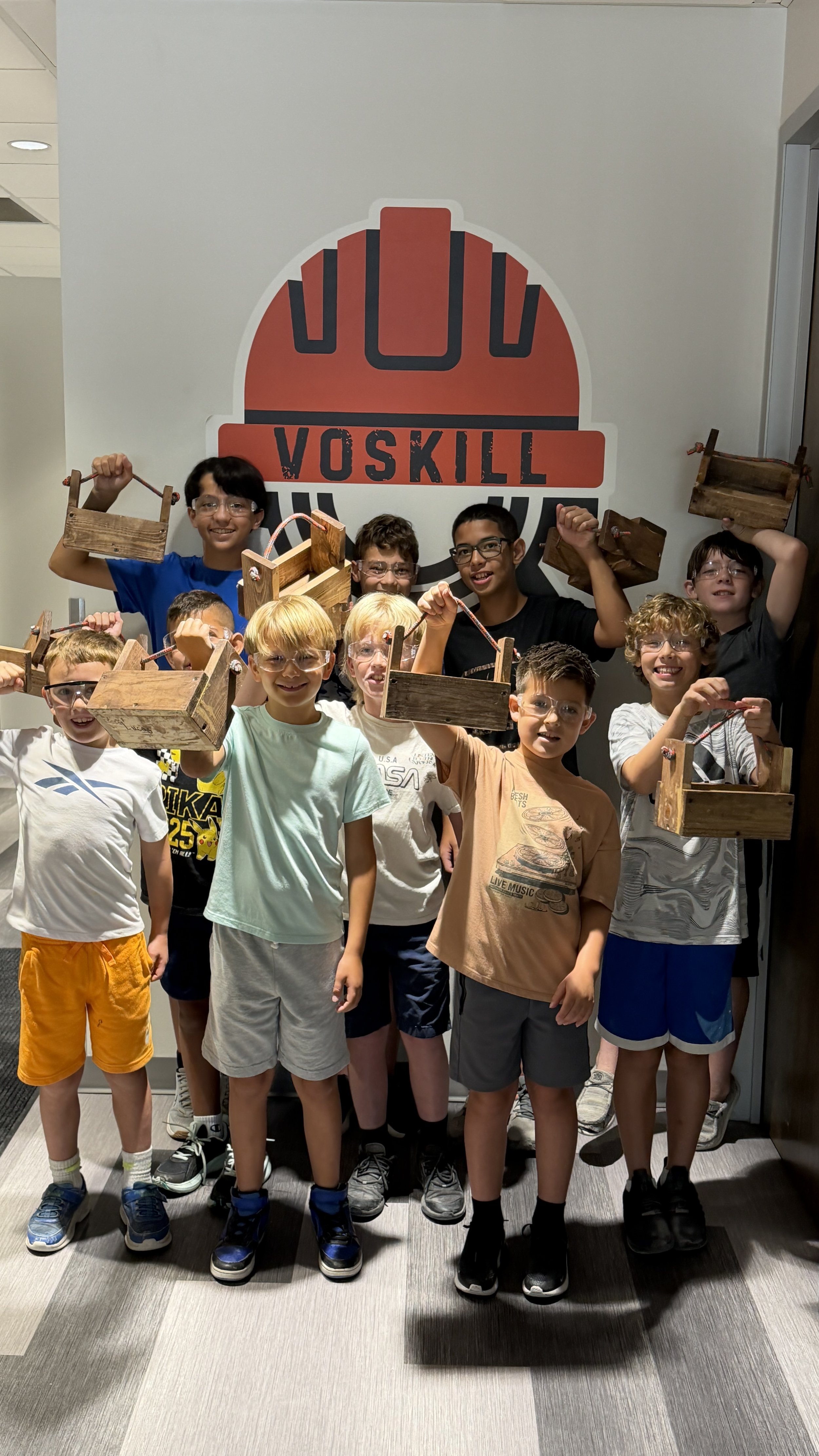 Group of children wearing safety glasses, holding wooden crates, smiling in front of a large Voskills logo
