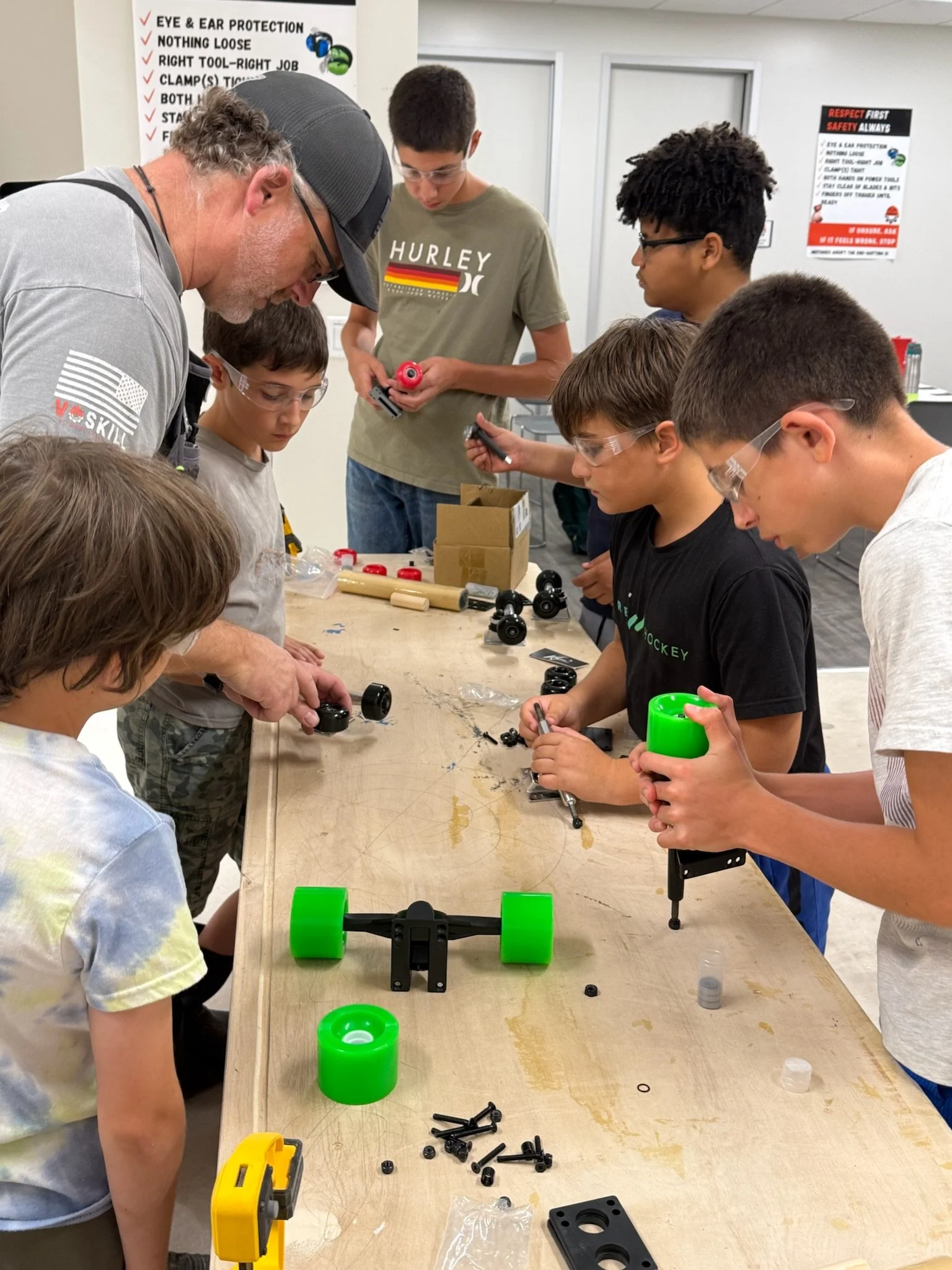 A group of children and an adult working on a robotics project at a workshop table with various machine parts and tools, in a classroom with informational posters on safety and protection on the wall.