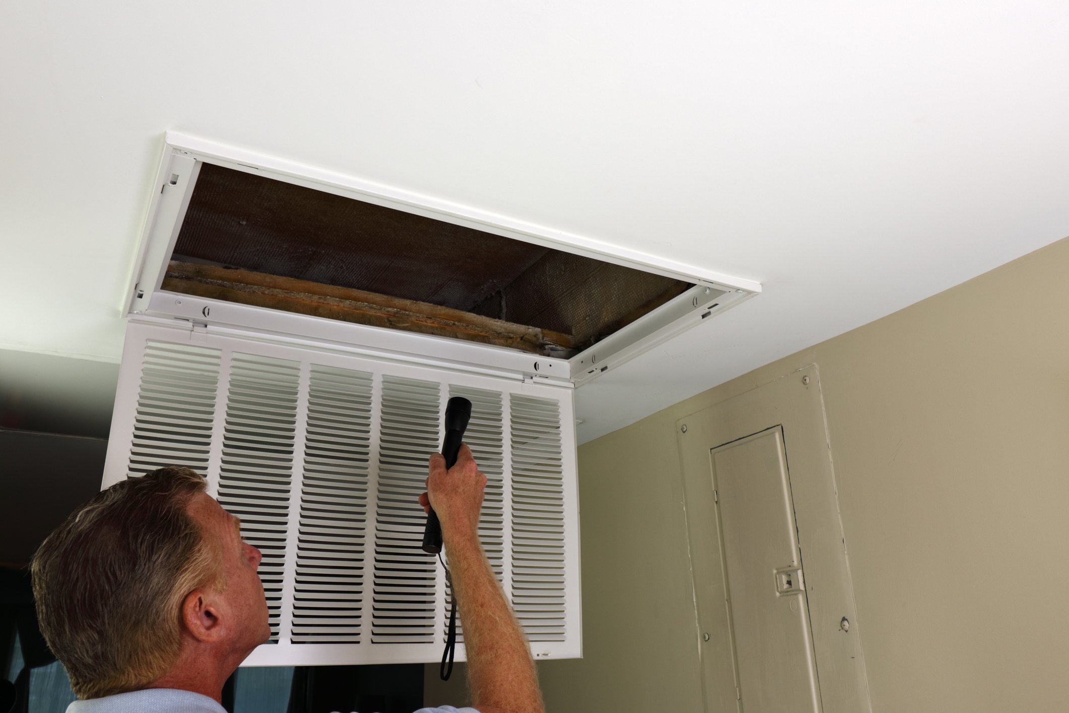 A man inspecting an open ceiling vent with a flashlight in a room.