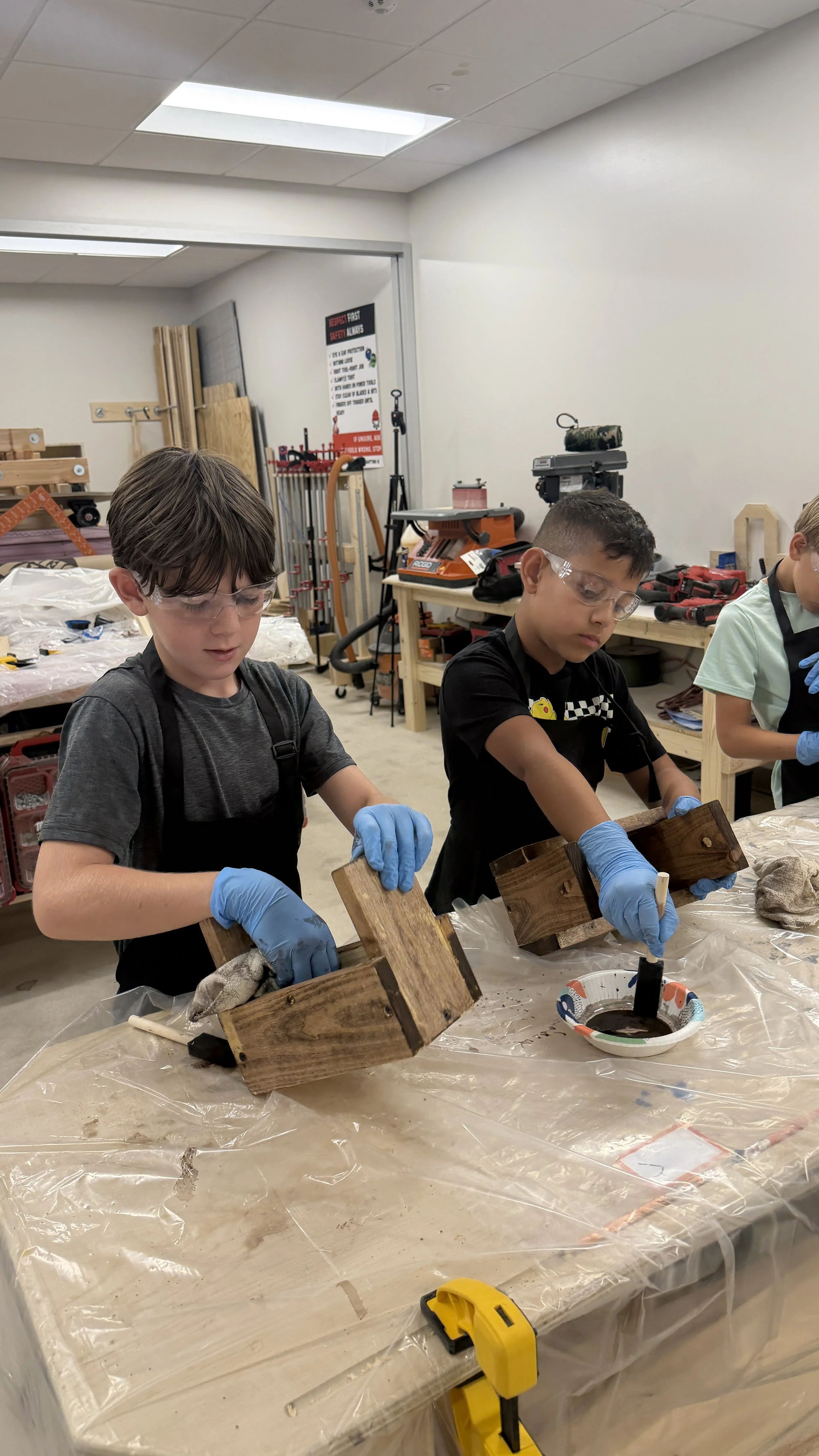 Two boys wearing safety goggles and gloves working on wooden projects in a woodworking classroom.