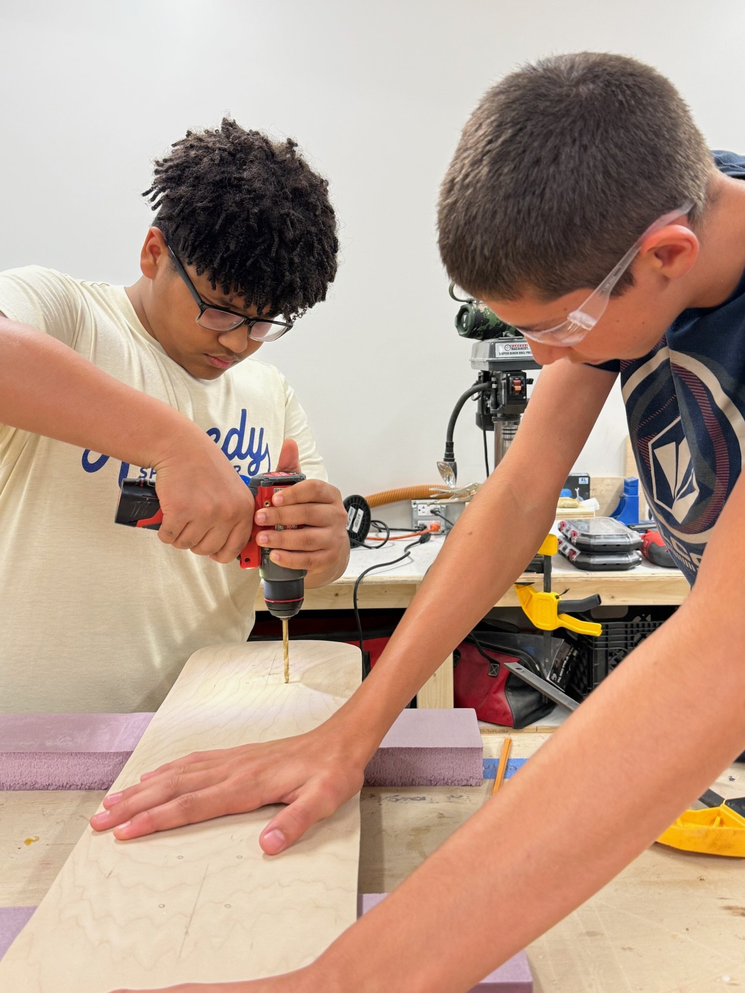 Two boys working on a woodworking project in a workshop, one using a drill, the other stabilizing a piece of wood.