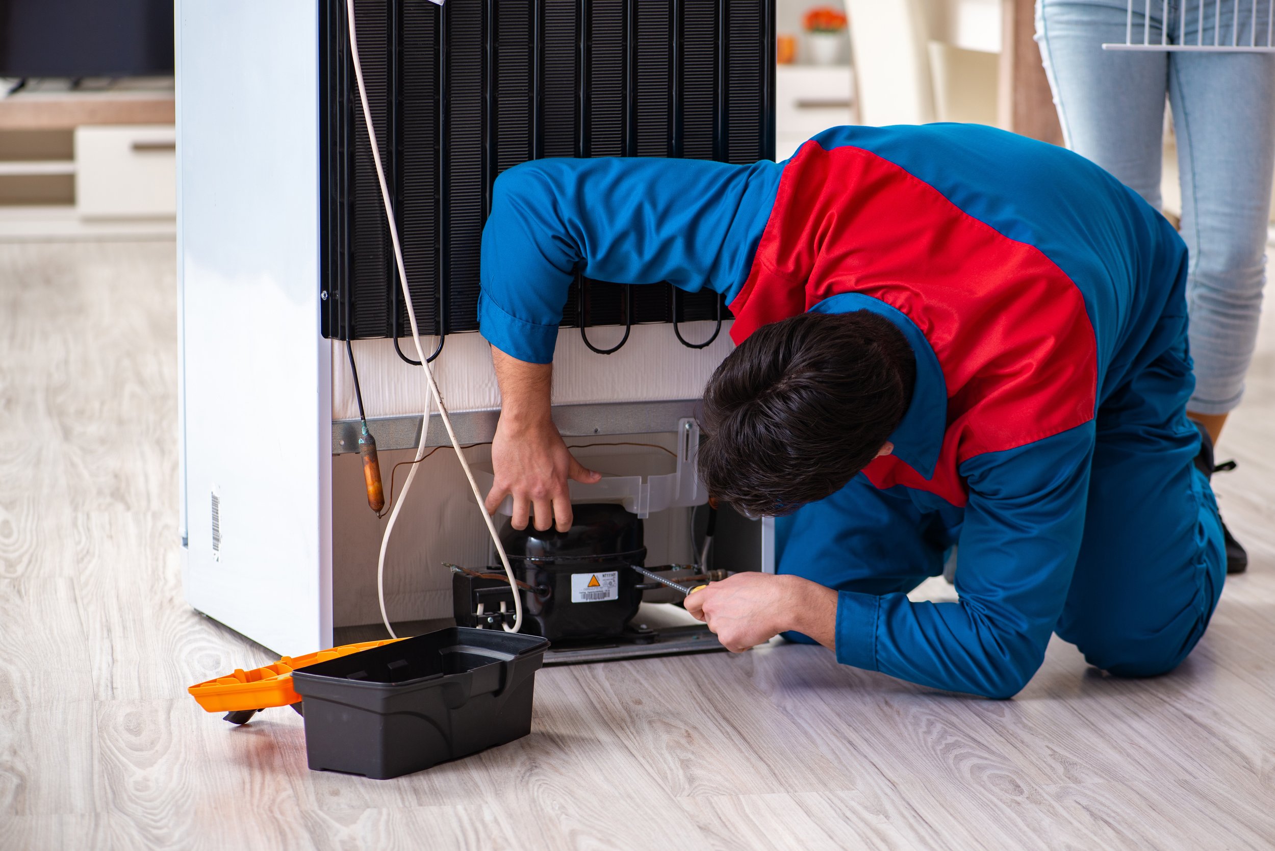A technician repairs a refrigerator while kneeling on the floor, with tools and a black toolbox nearby.