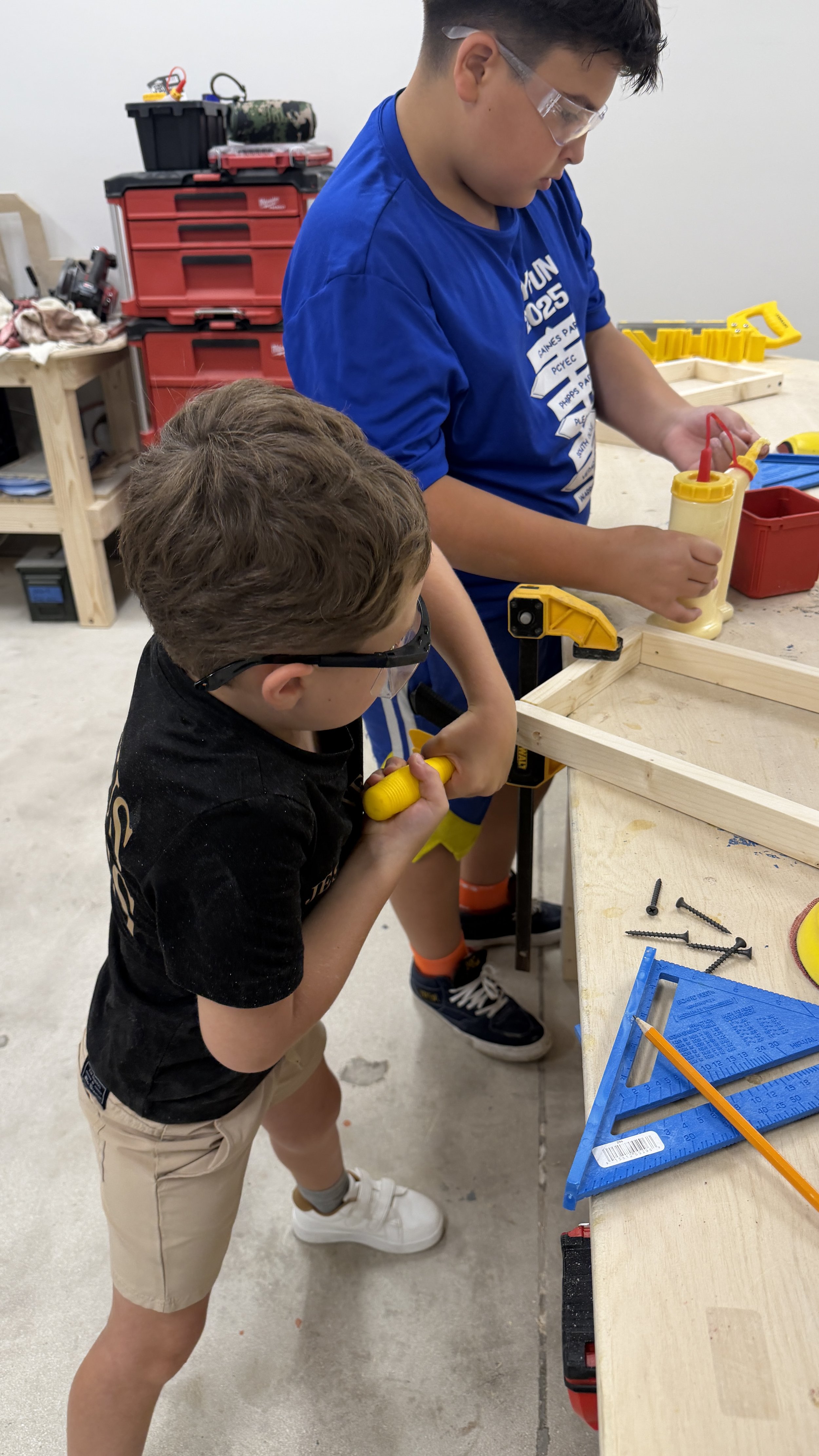 Two young boys are working together on a woodworking project at a workshop table. One boy is holding a yellow screwdriver, and the other boy is holding a bottle of wood glue. They are surrounded by tools and materials, with a red and black tool chest in the background.