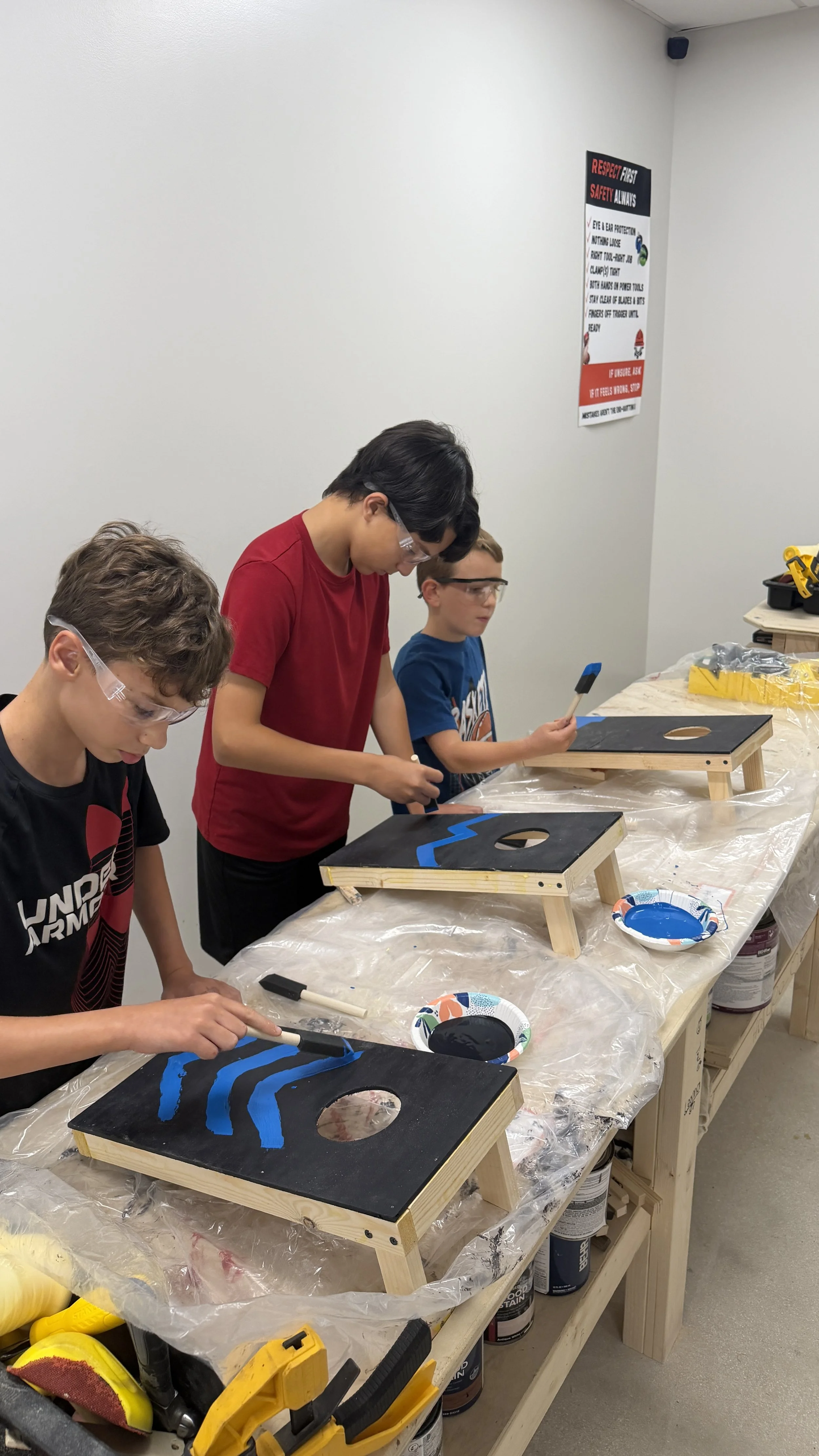 Four boys painting on black wooden boards with blue paint in a classroom or workshop setting, with various painting supplies and tools on the table.