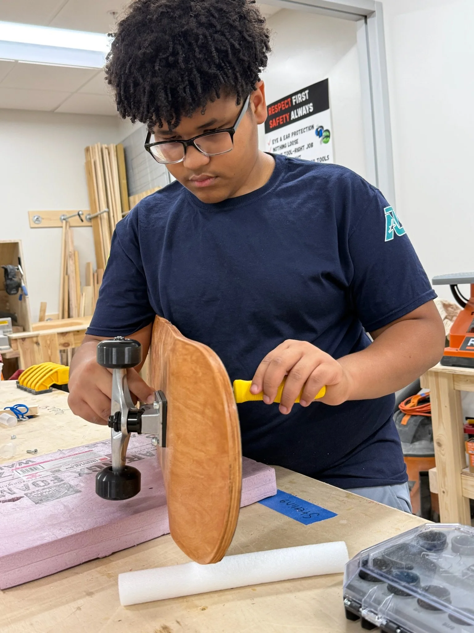 A young person with glasses and curly hair working on a skateboard in a woodworking shop. The person is fastening its wheels with a wrench over a workbench with tools and foam.