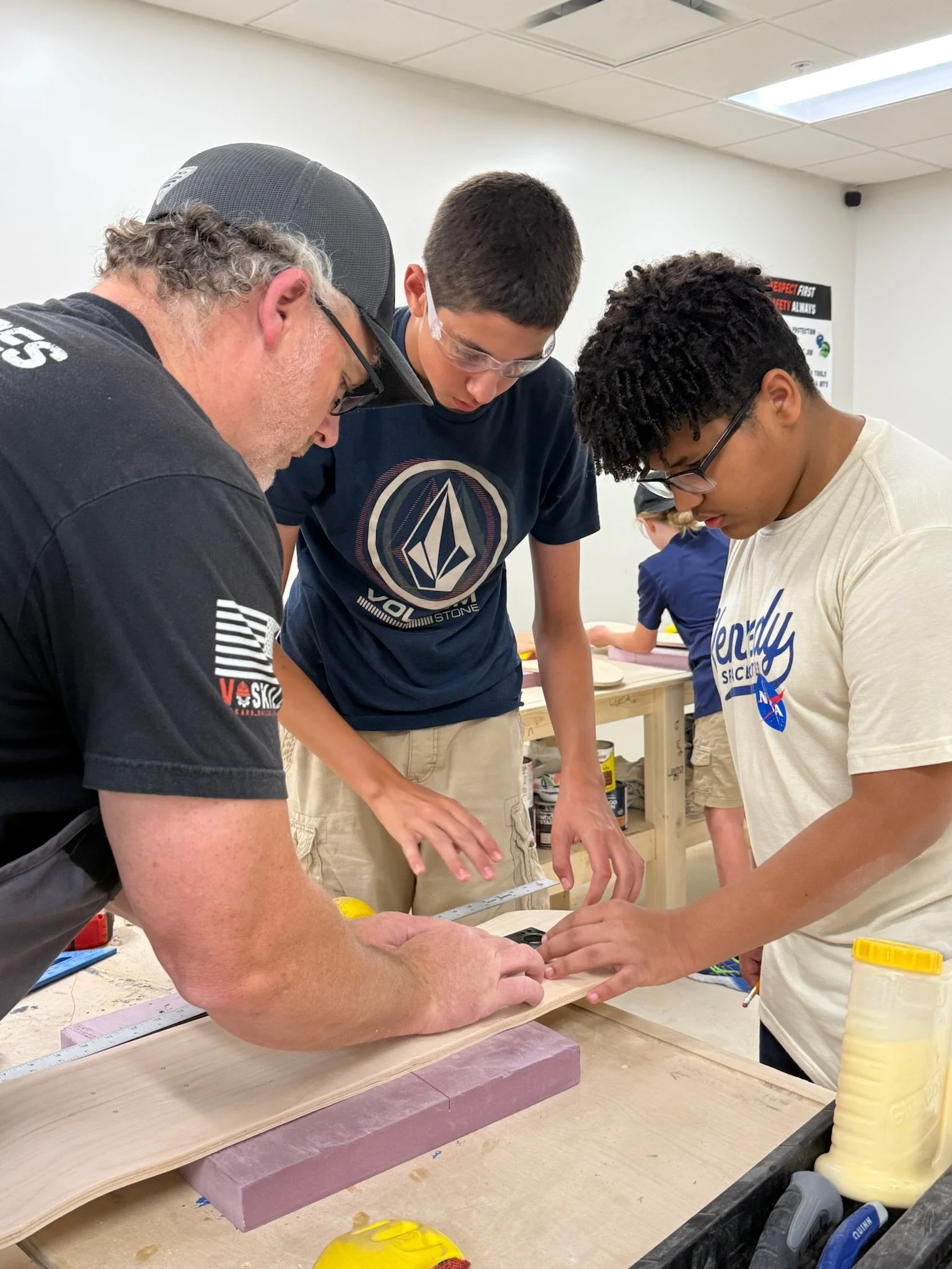 Three people working on a woodworking project in a workshop. They are measuring and examining a piece of wood on a workbench, with tools and supplies around them.