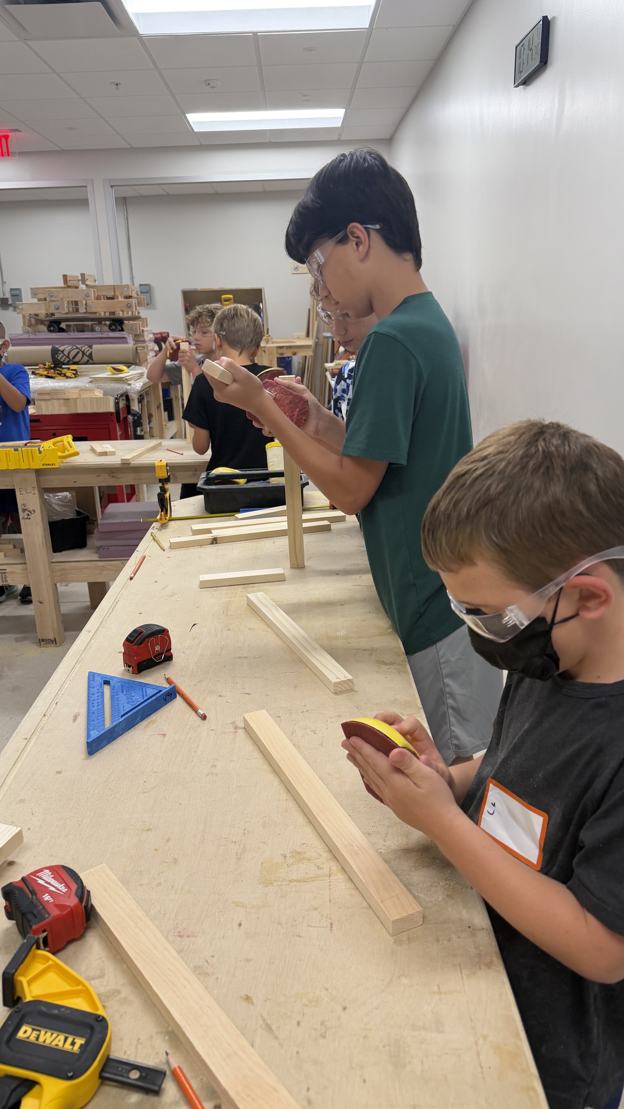 Group of children in a workshop, working on woodworking projects with tools and wooden blocks on a long work table. Some kids are sanding or measuring wood pieces, wearing safety goggles and masks.