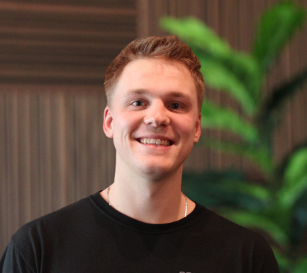 A young man with short light brown hair smiling, wearing a black shirt, with green plants and a wooden background behind him.