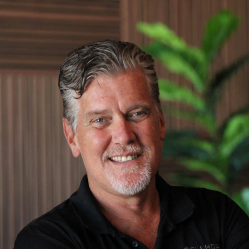 A middle-aged man with gray hair and a beard smiling at the camera, with green plants and wood-paneled walls in the background.