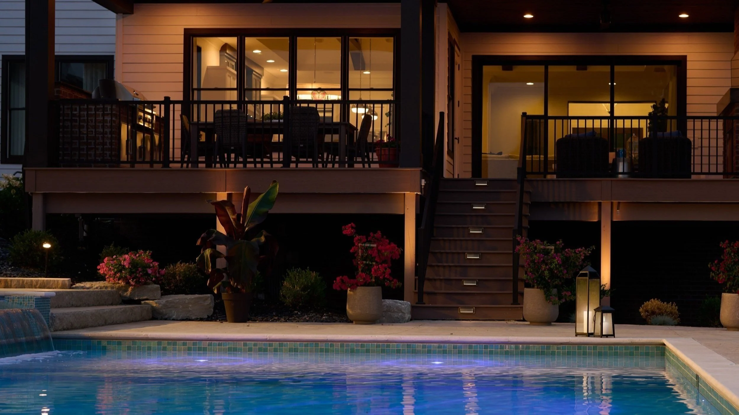 A nighttime view of a backyard with a swimming pool, a deck with outdoor furniture, potted plants, and lanterns.