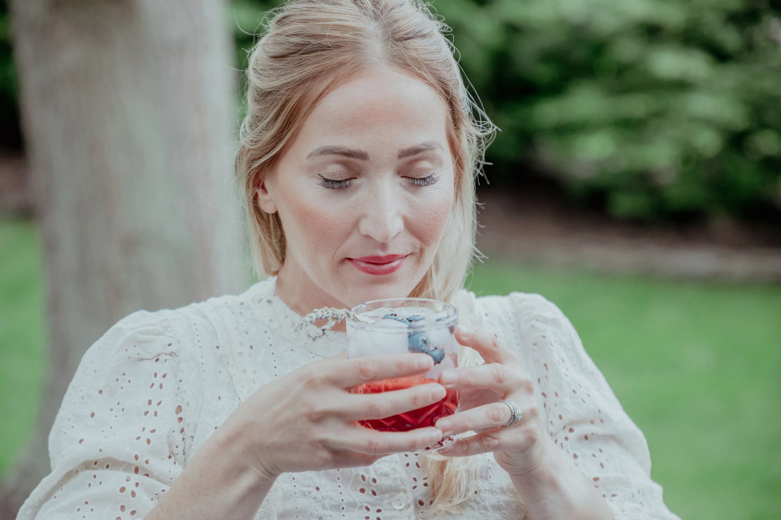 A woman with light blonde hair and wearing a white eyelet blouse holds a glass of red and clear layered beverage, possibly with ice and berries, outdoors near a tree.