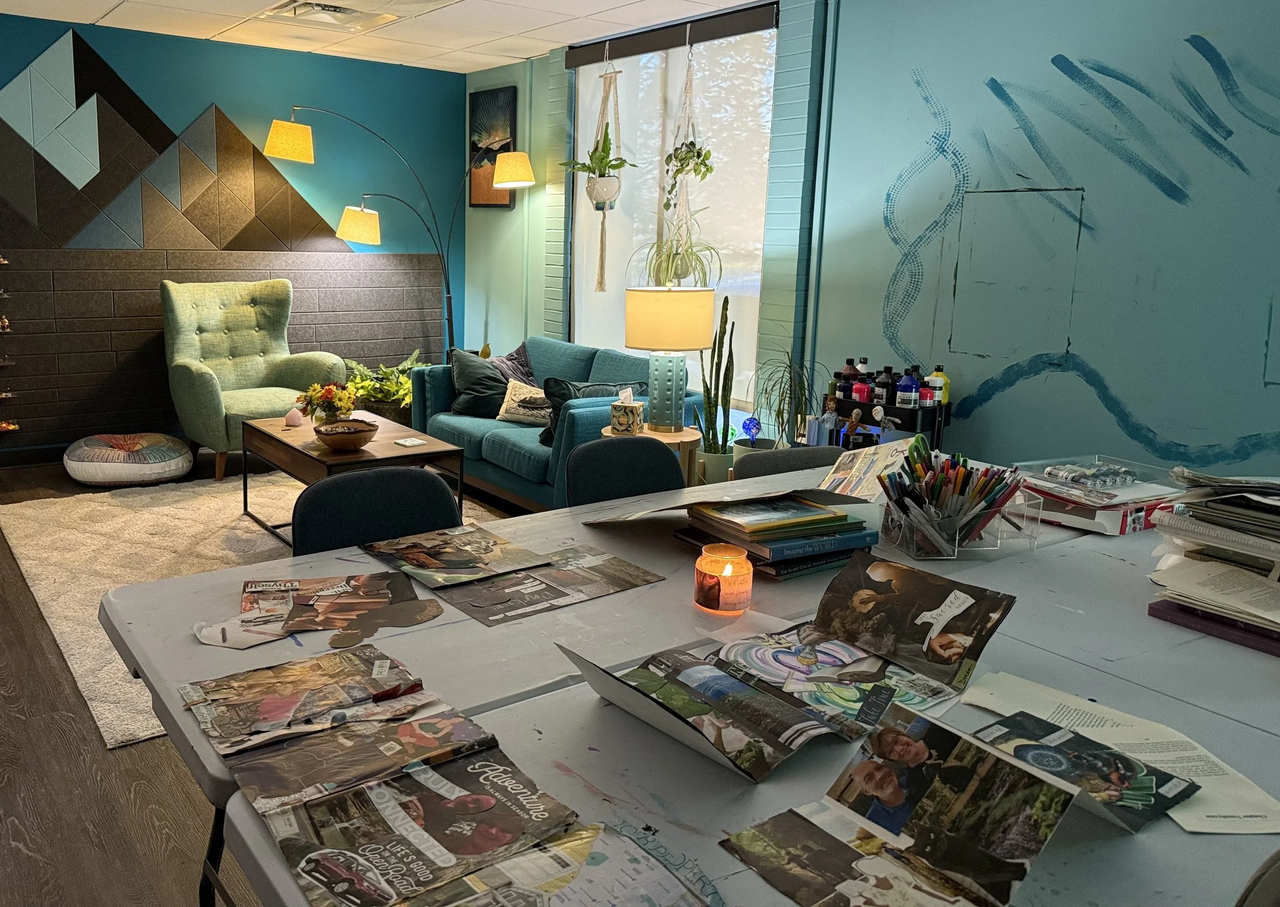 Indoor art studio office space with teal and black walls, a green armchair, a teal sofa, floor lamp, hanging plants, and a window with blinds. There are tables with books and magazines, collages, a candle and an art piece on the wall, a calm space.