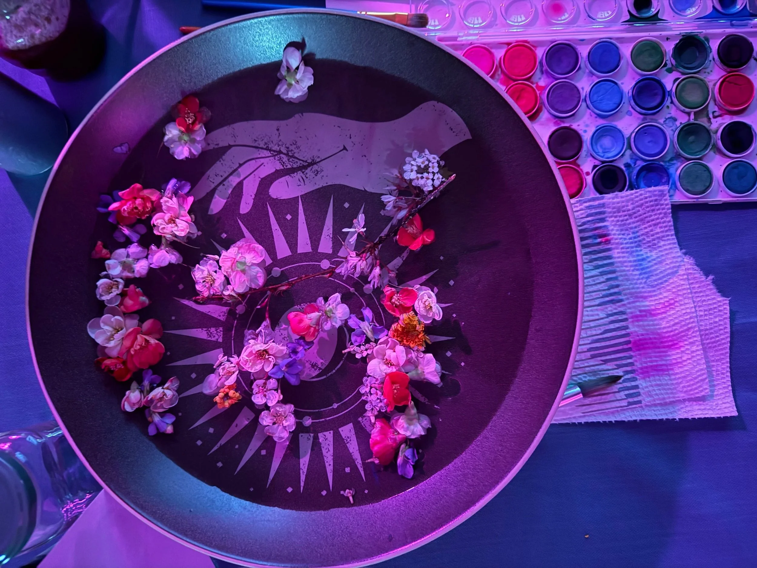 A round black bowl filled with water with white graphic of a hand holding a flower, surrounded by pink and white flowers. To the right, there is a tray of colorful watercolors and a textured white towel, all illuminated by purple and pink lighting.