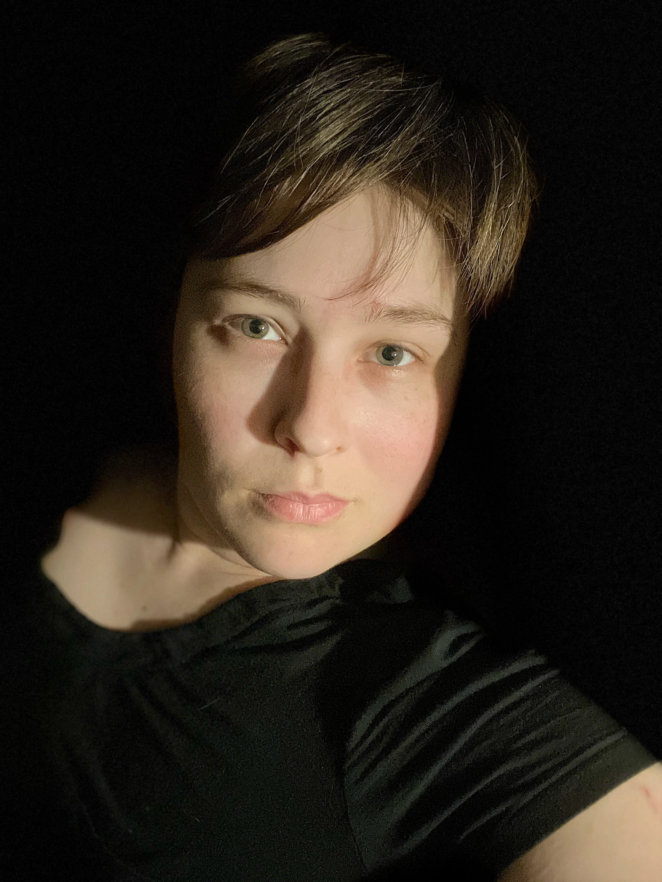 A young person with short brown hair, green eyes, and light skin, wearing a black top, looking directly at the camera with a neutral expression.