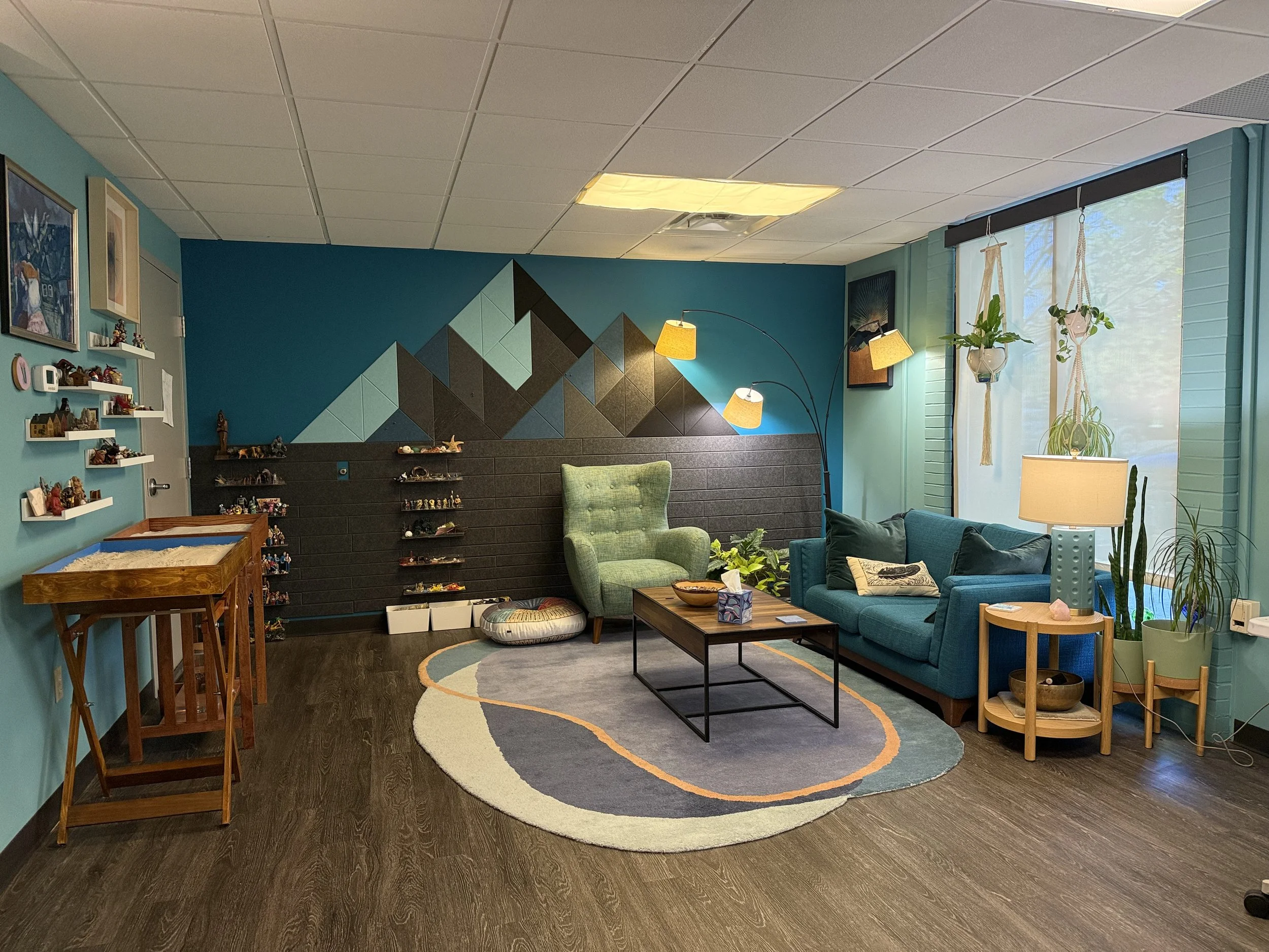 A cozy art studio office room with teal walls, a geometric mountain mural, shelves with small figurines, a green armchair, a blue sofa, a wooden coffee table, floor lamps with yellow shades, and large windows with hanging planters and blinds.