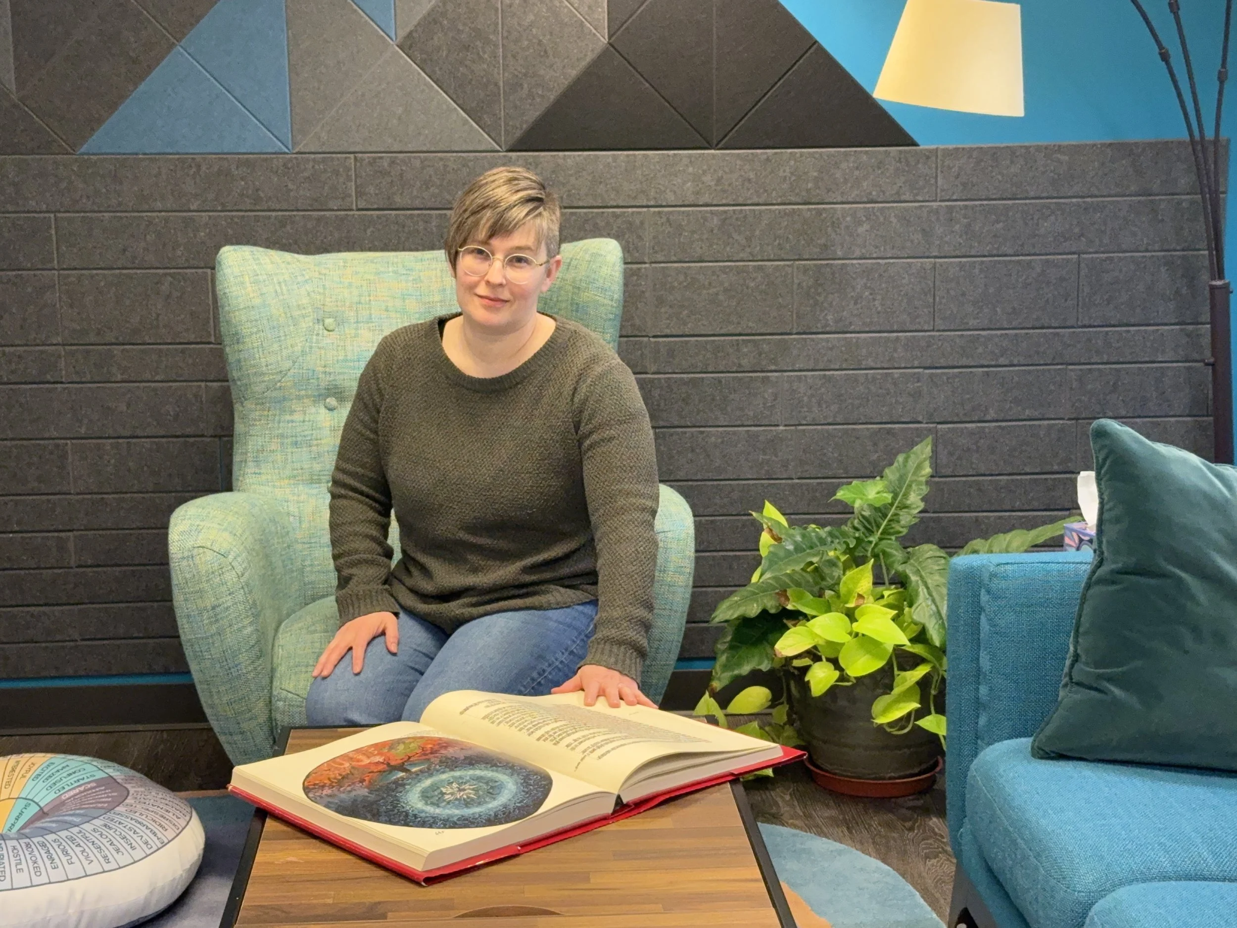 A woman with short hair, glasses, and an olive sweater sitting on a green armchair in a modern indoor space with a black brick wall background, the Red Book open with colorful painting by Jung and text on a table in front of her, and plants nearby.