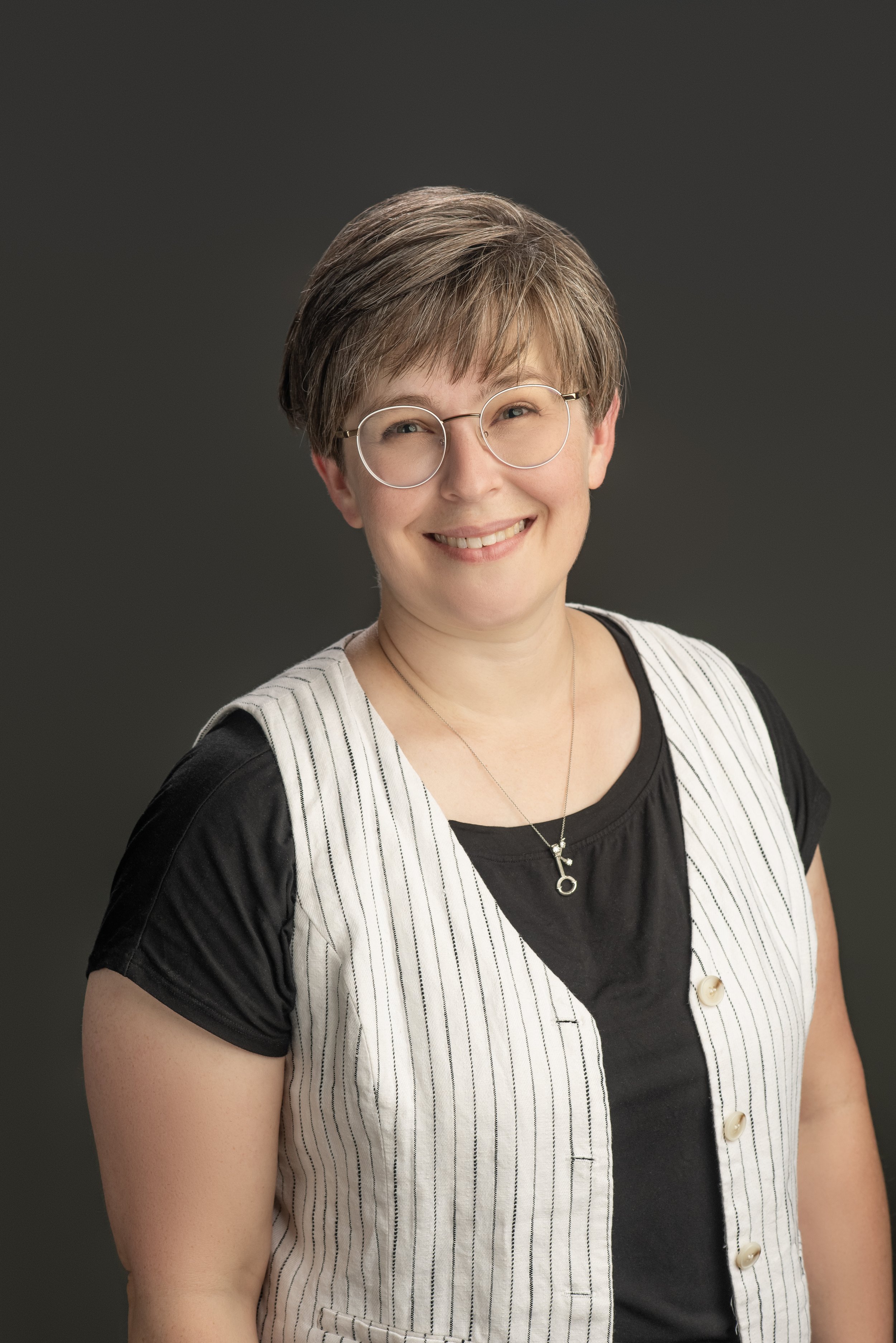 Portrait of a smiling woman with glasses, short brown hair, wearing a black and white striped vest over a black shirt, against a dark background.