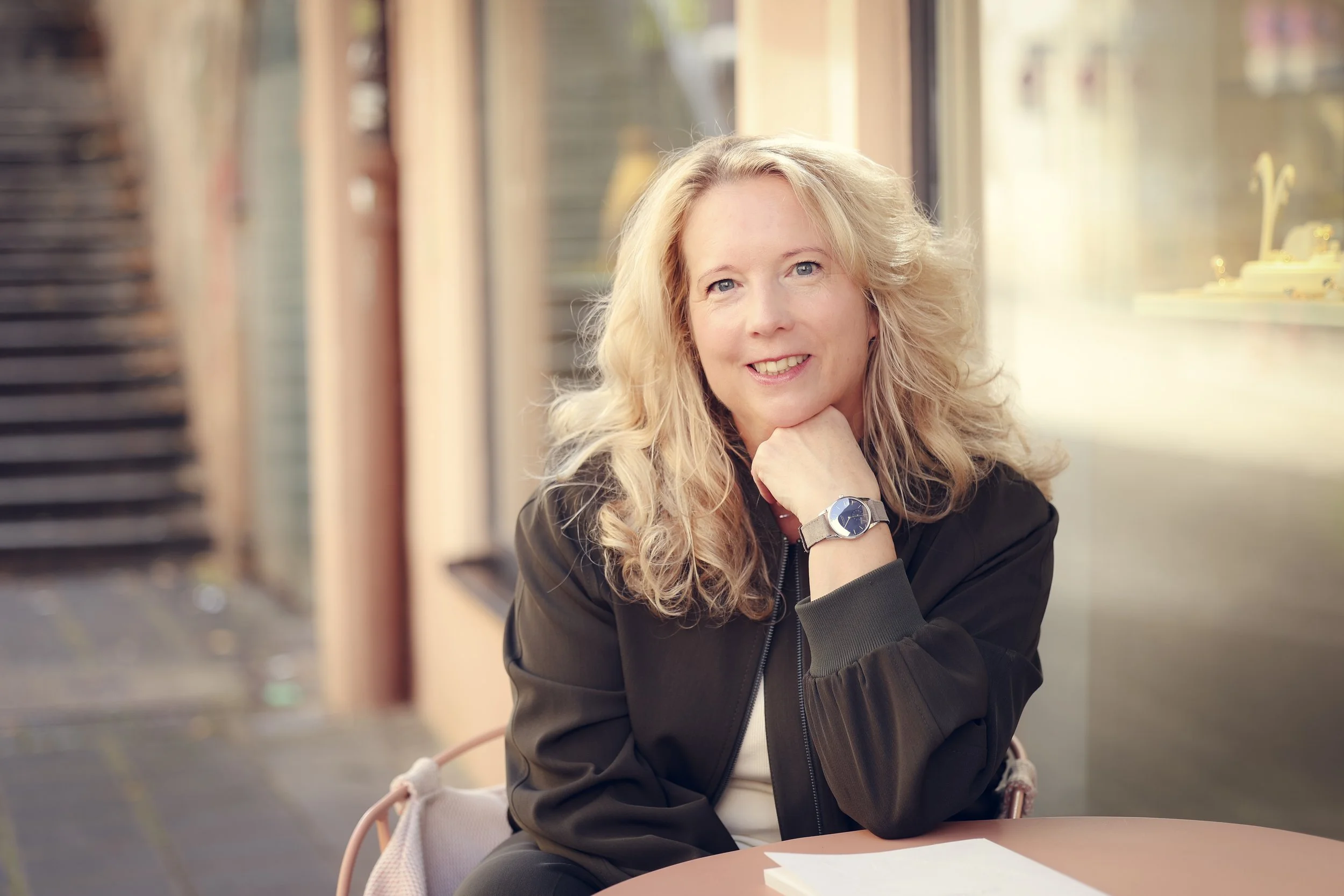 Eine lächelnde Frau, die Gründerin von taxculture, mit blonden, lockigen Haaren sitzt an einem Tisch in einem Café, trägt eine schwarze Jacke und eine Armbanduhr.