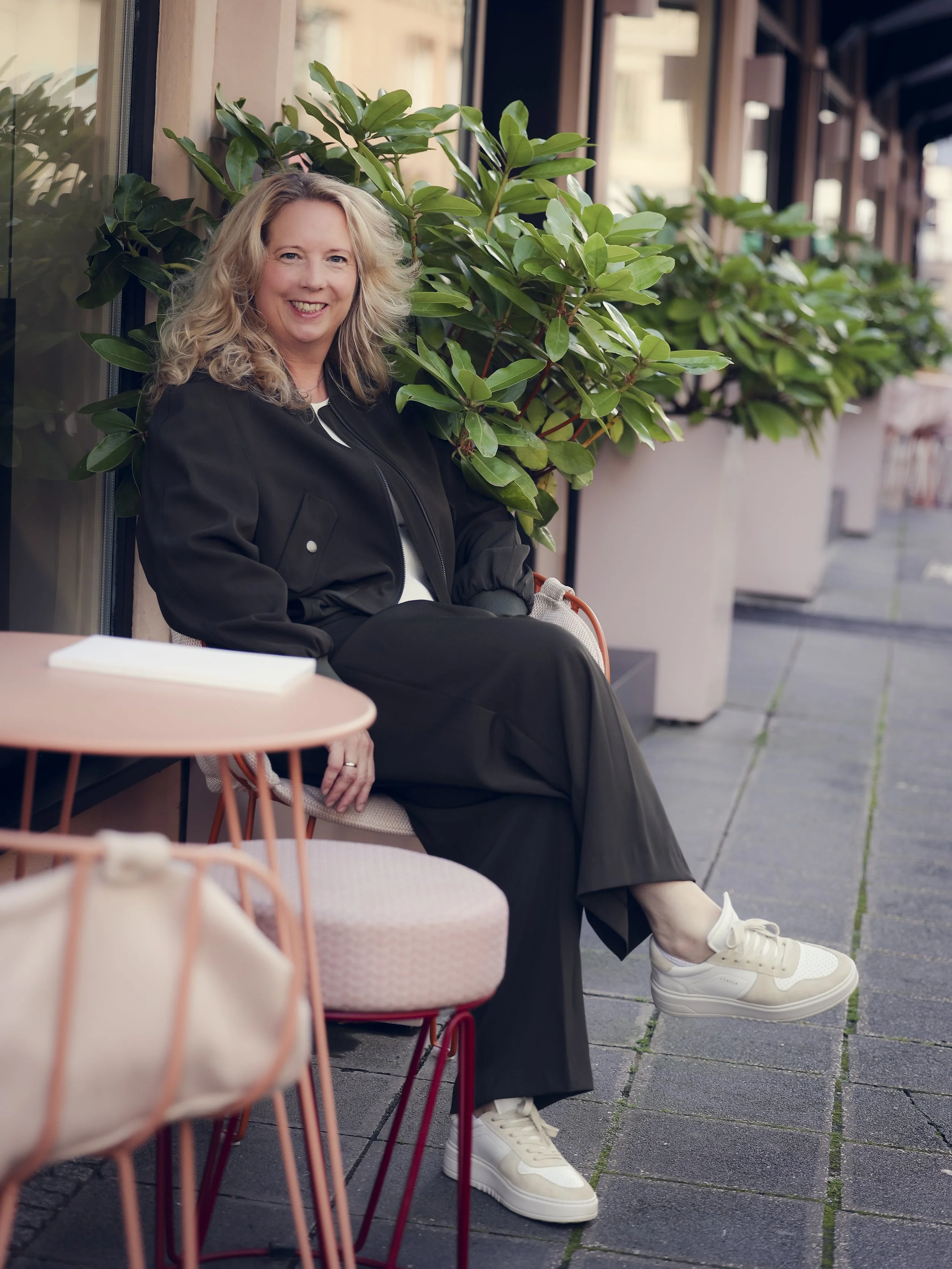 Die Gründerin von taxuclture mit lockigem blonden Haar sitzt lächelnd auf einem stromlinienförmigen Stuhl im Freien neben einem kleinen runden Tisch, umgeben von großen grünen Pflanzen, vor einem Café in einer Stadtstraße.