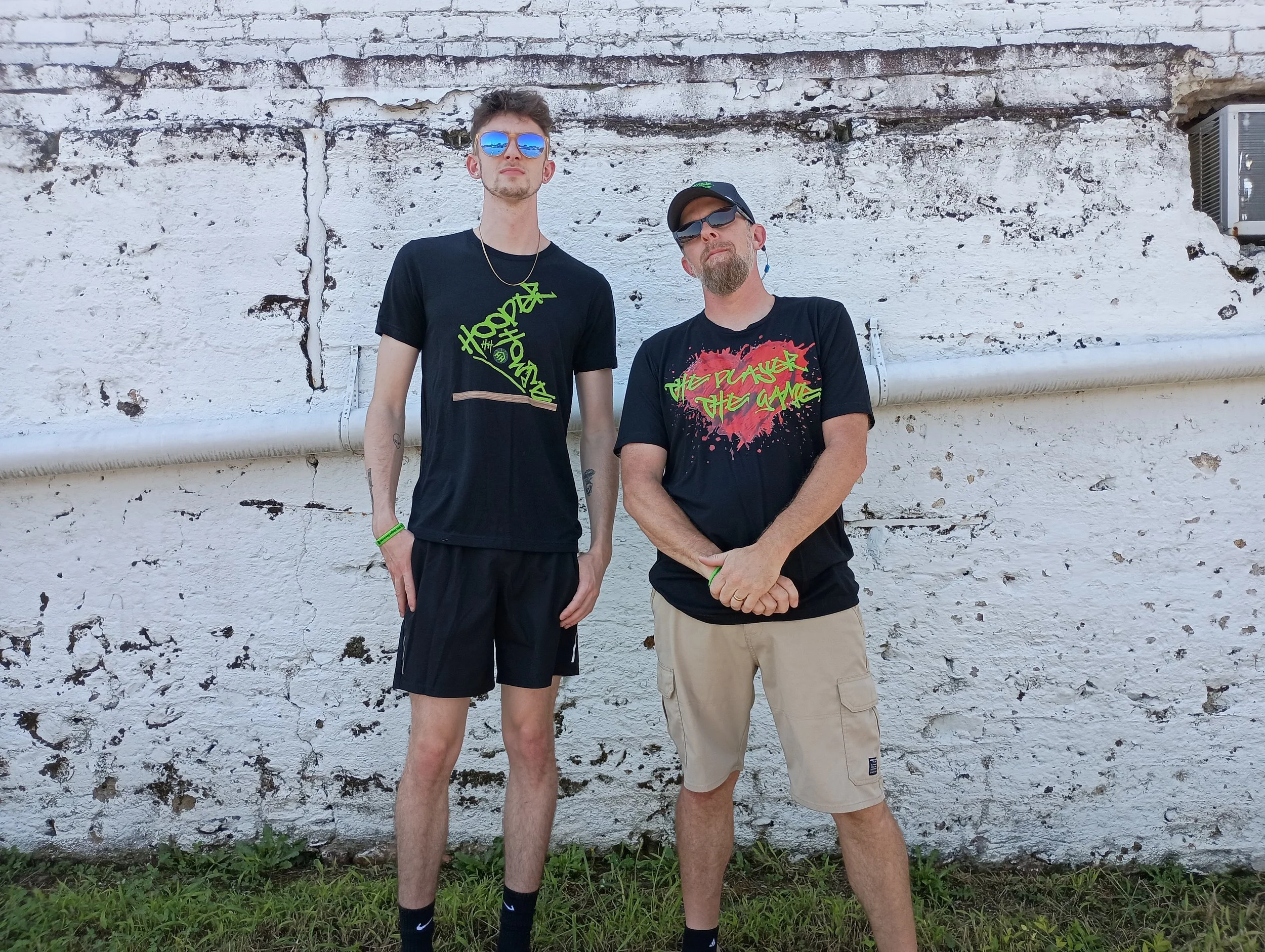 Two men standing outdoors in front of a white brick wall with a pipe. They are wearing casual black T-shirts and shorts, sunglasses, and accessories like bracelets and necklaces. One man has short, dark hair, and the other has a beard and is wearing a cap.