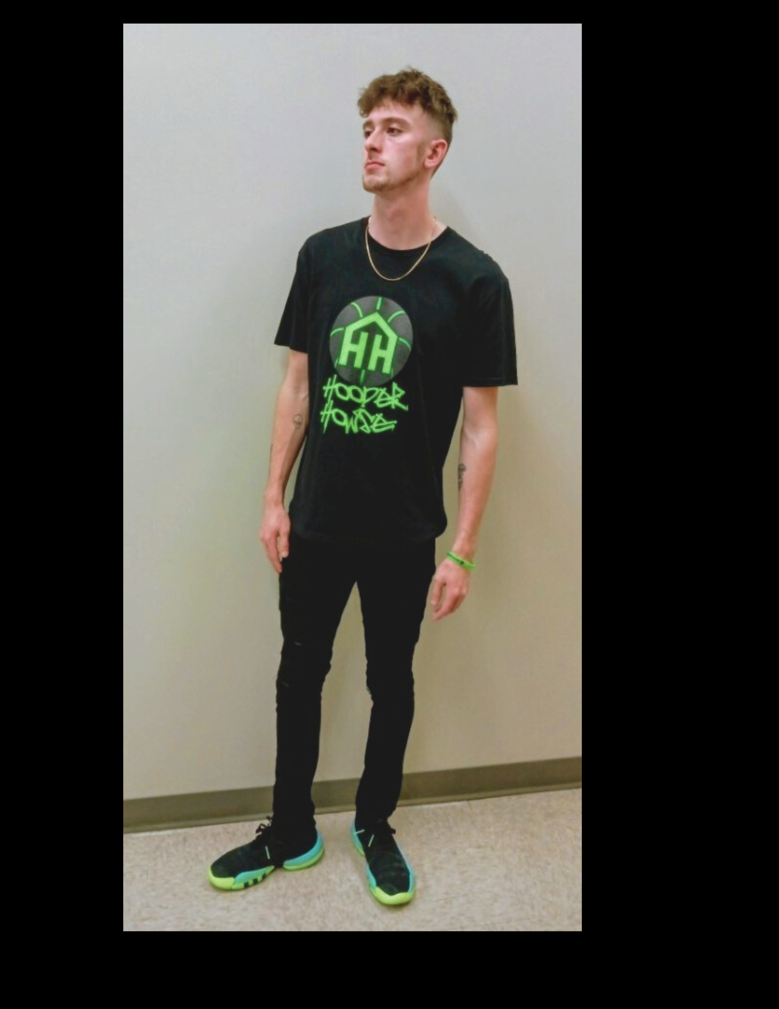 A young man in black sneakers with neon green soles, black skinny jeans, a black t-shirt with green graphics, standing against a plain gray wall.