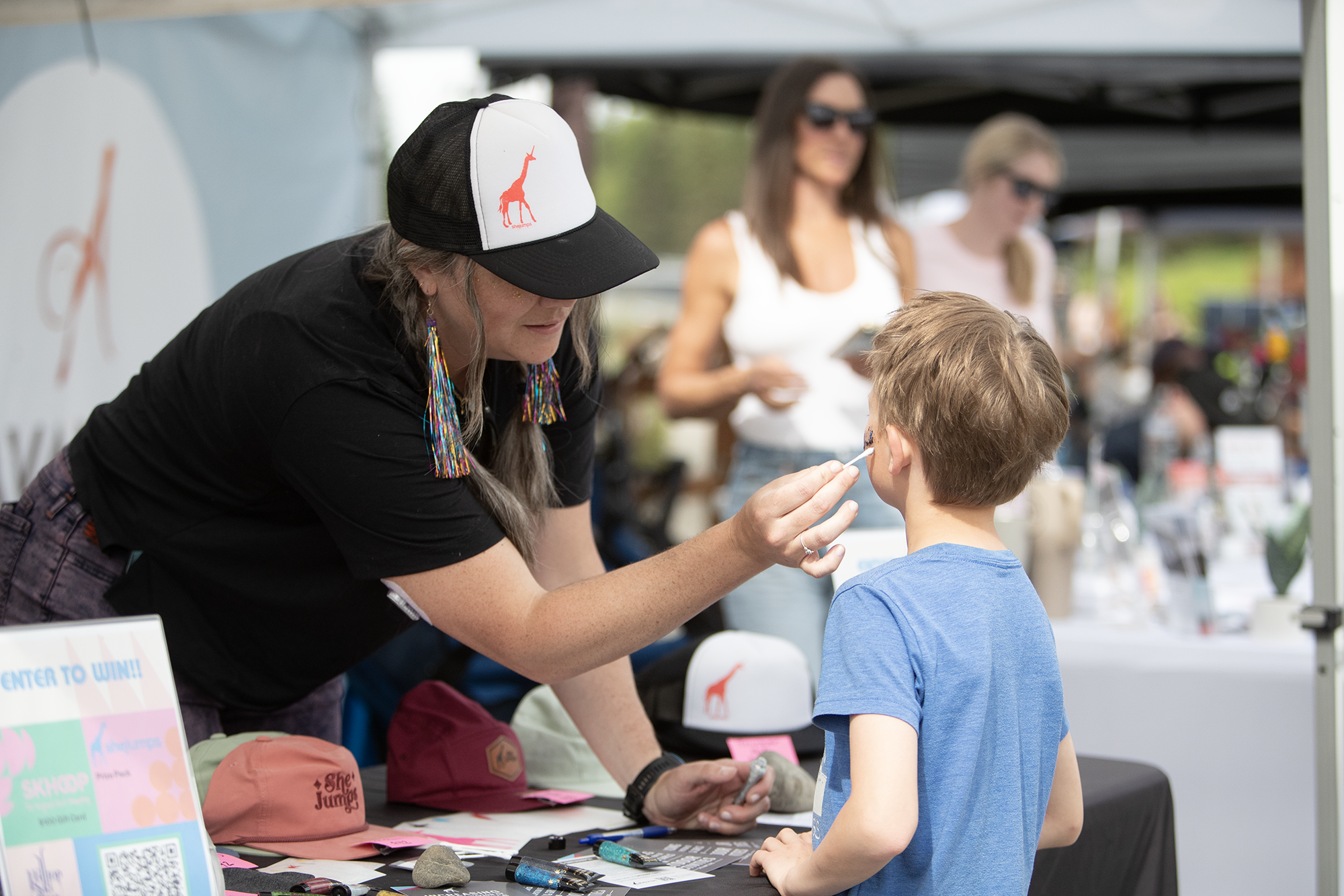 A woman wearing a black cap with a giraffe patch and colorful earrings is giving a medical check to a young boy at an outdoor event. Two women are blurred in the background under tents, and there are caps and promotional materials on the table.