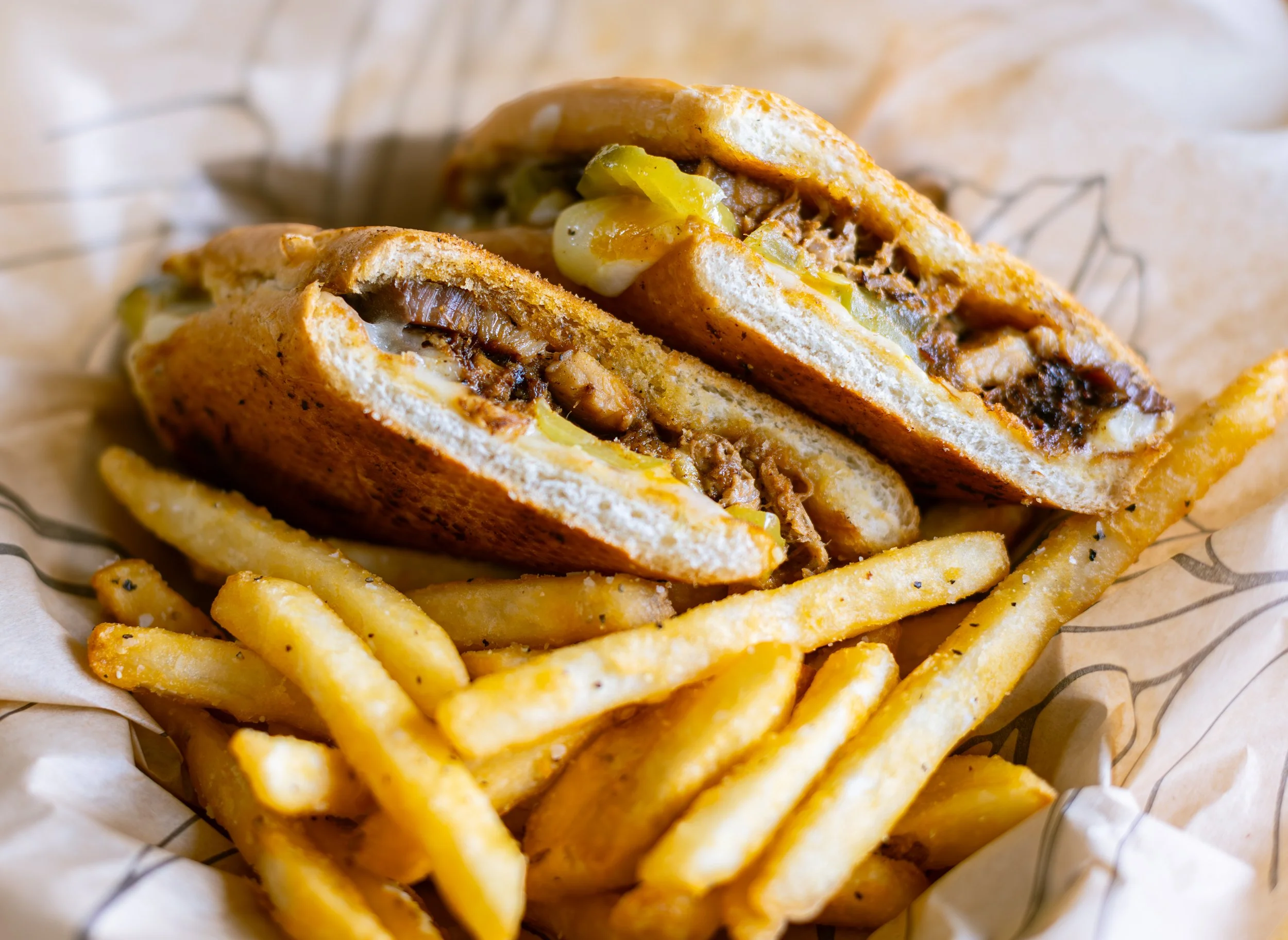 A close-up image of a cheeseburger with pickles and melted cheese, served with French fries in a basket lined with paper.