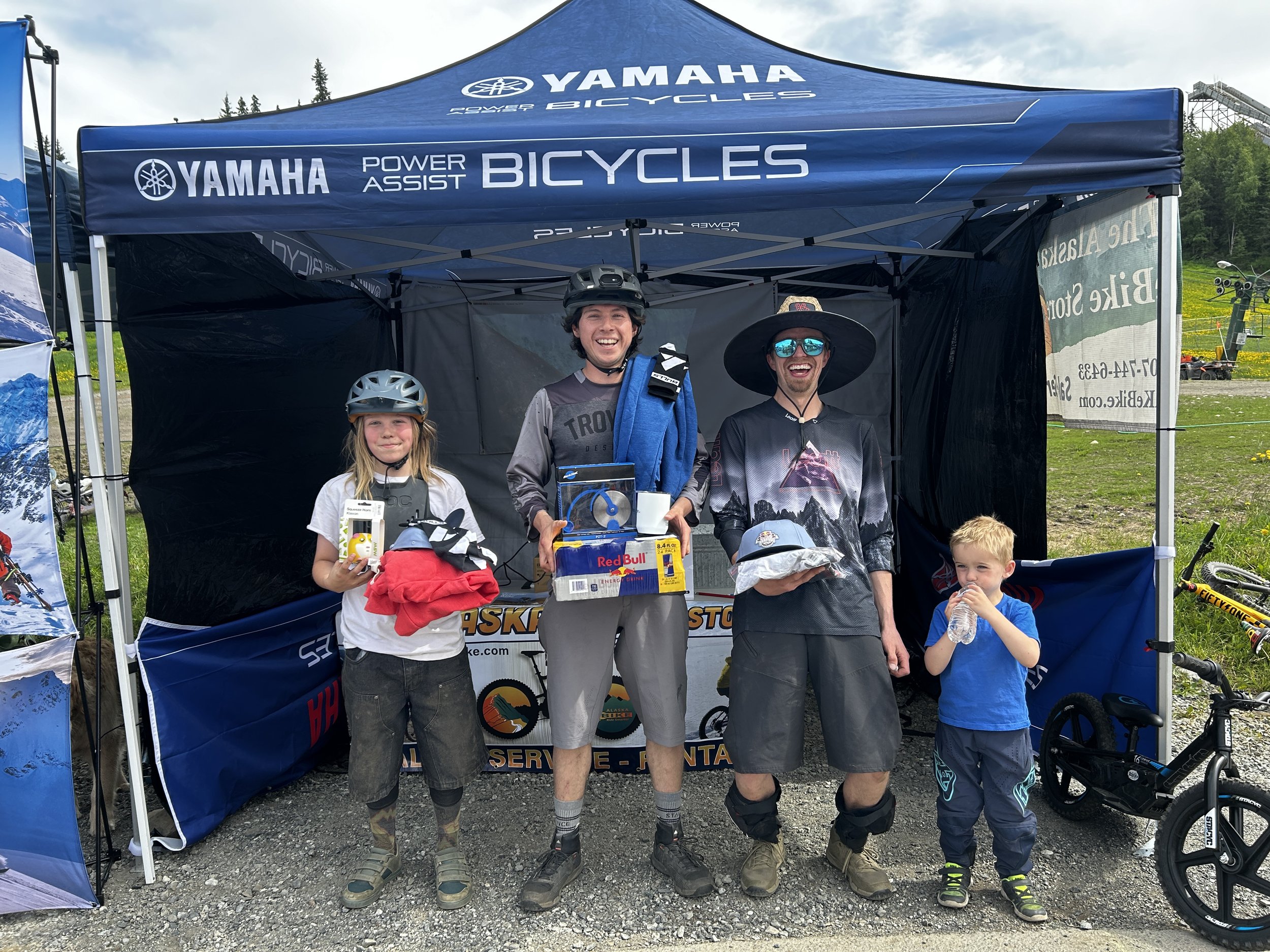 Four smiling people, including three adults and a child, standing under a blue Yamaha Powers Assist Bicycles tent, holding awards and prizes, at an outdoor event with bikes and green landscape in the background.