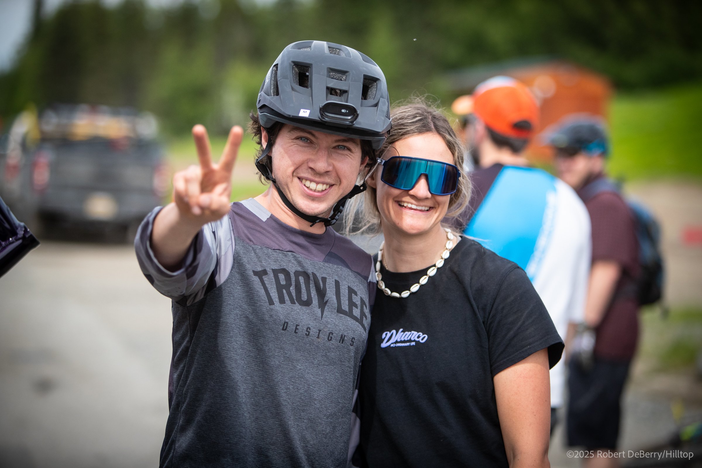 A smiling young man in a bike helmet making a peace sign, standing next to a smiling woman with sunglasses at an outdoor event.
