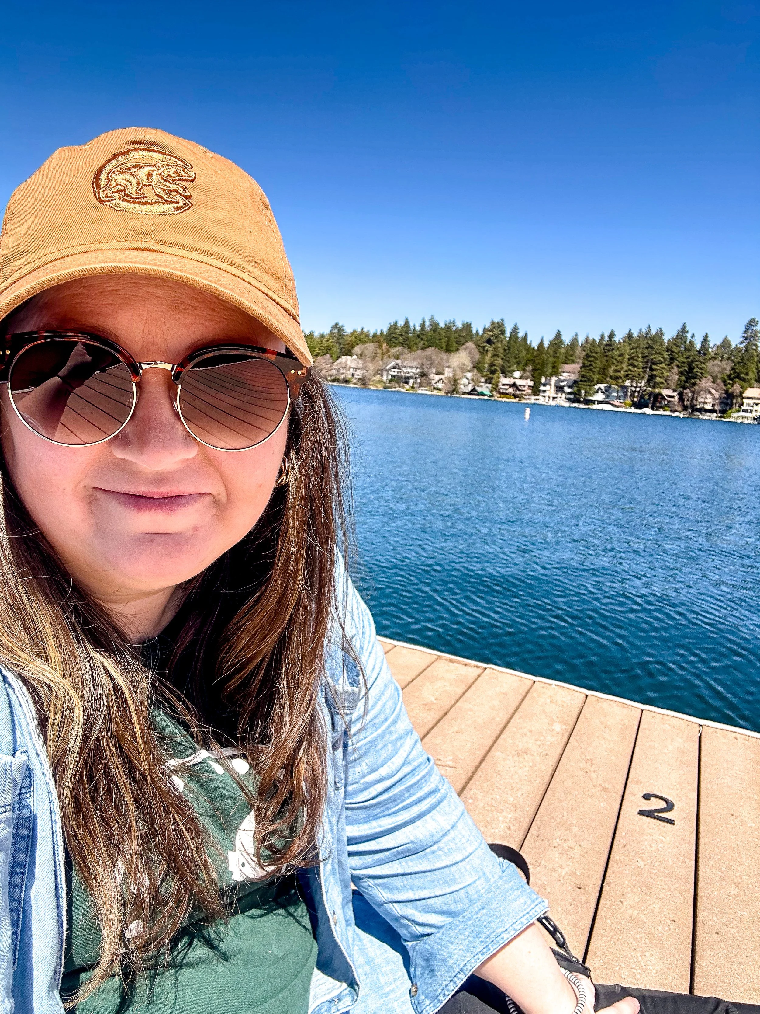 A woman wearing sunglasses and a tan cap takes a selfie by a lake on a sunny day with houses and trees in the background.
