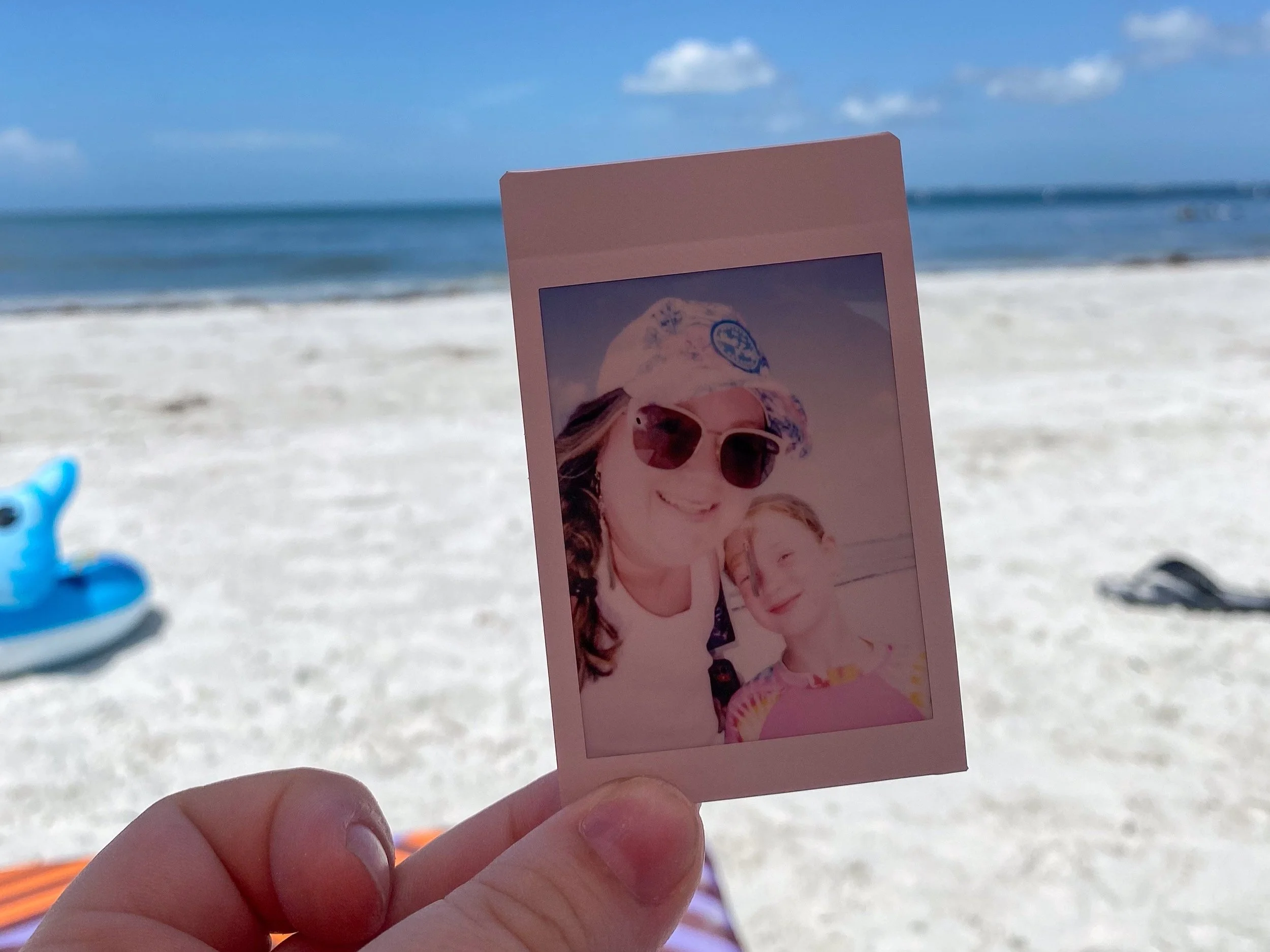 A hand holding a photograph of a smiling woman wearing sunglasses and a hat at the beach, with the ocean and a blue sky in the background.