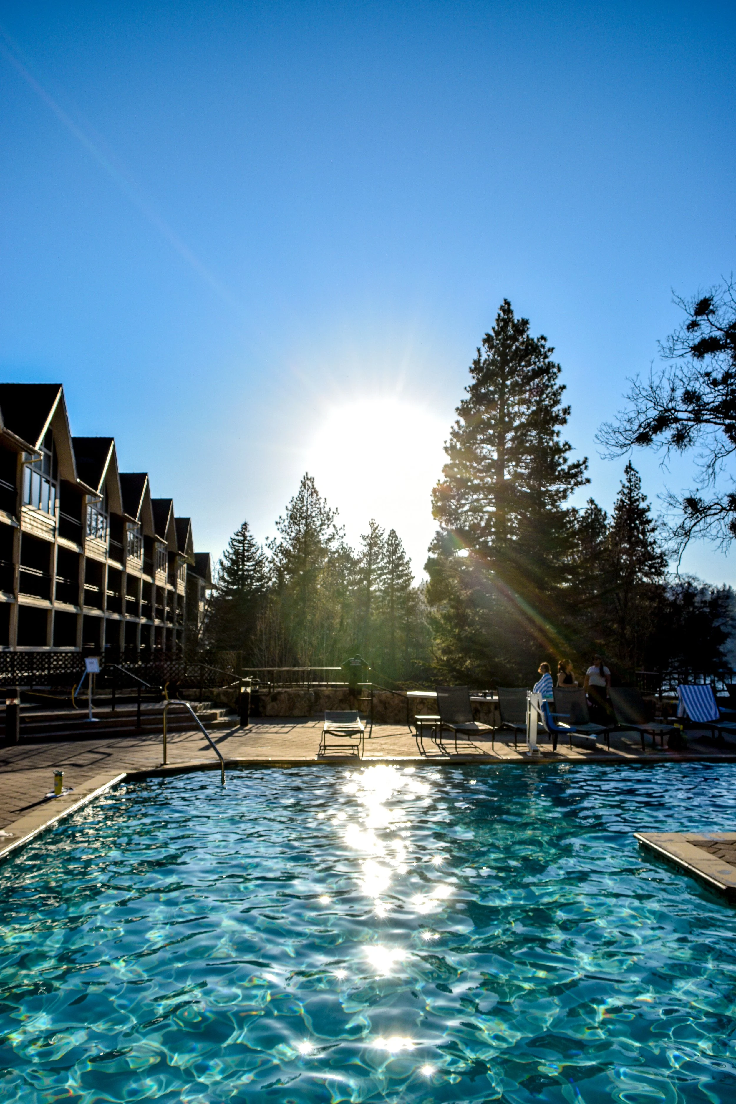 Swimming pool with lounge chairs and trees in the background, sunlight shining through the sky.