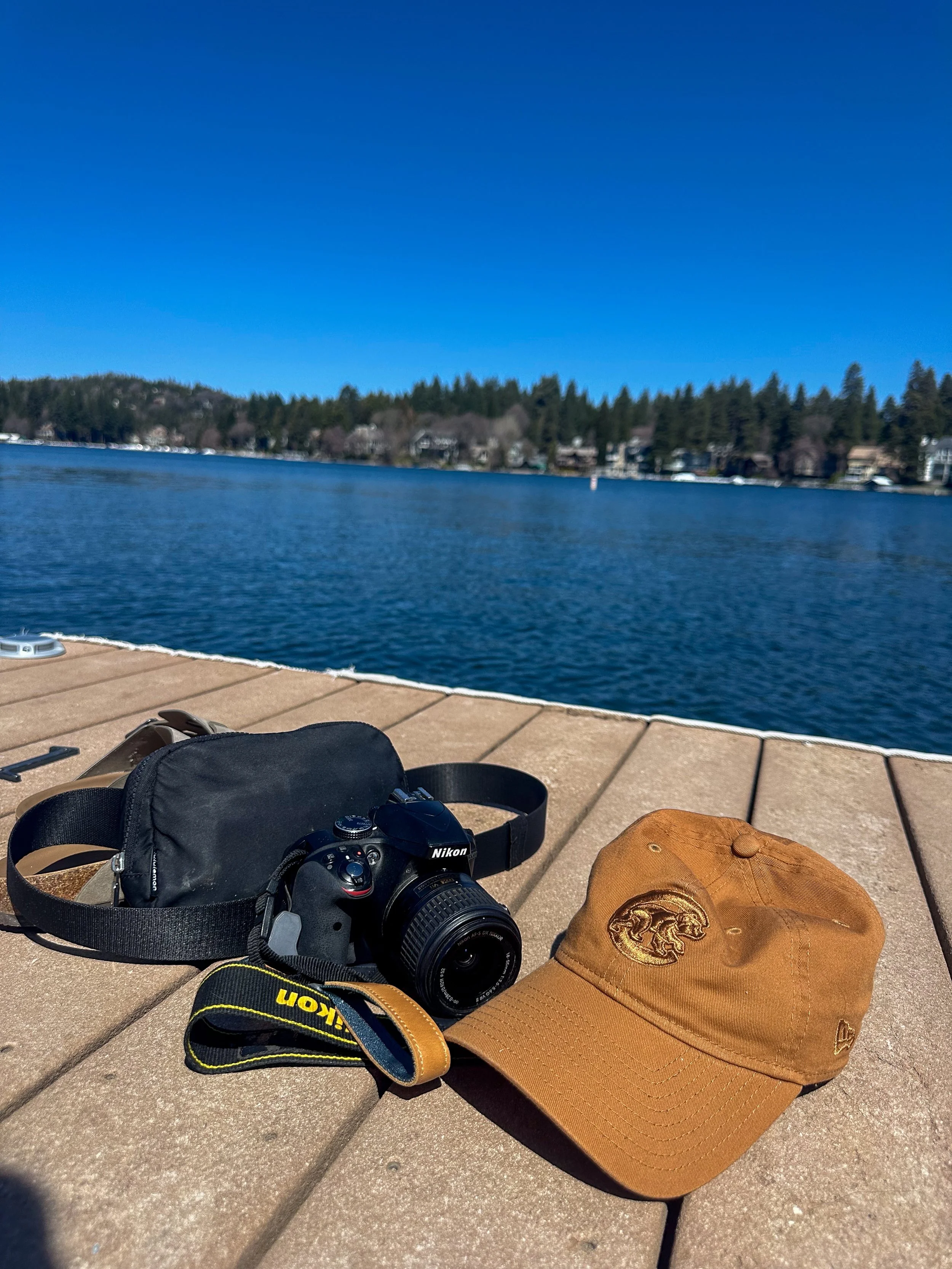 Camera, brown cap, and camera bag placed on wooden dock by a lake with clear blue sky and trees in the background.
