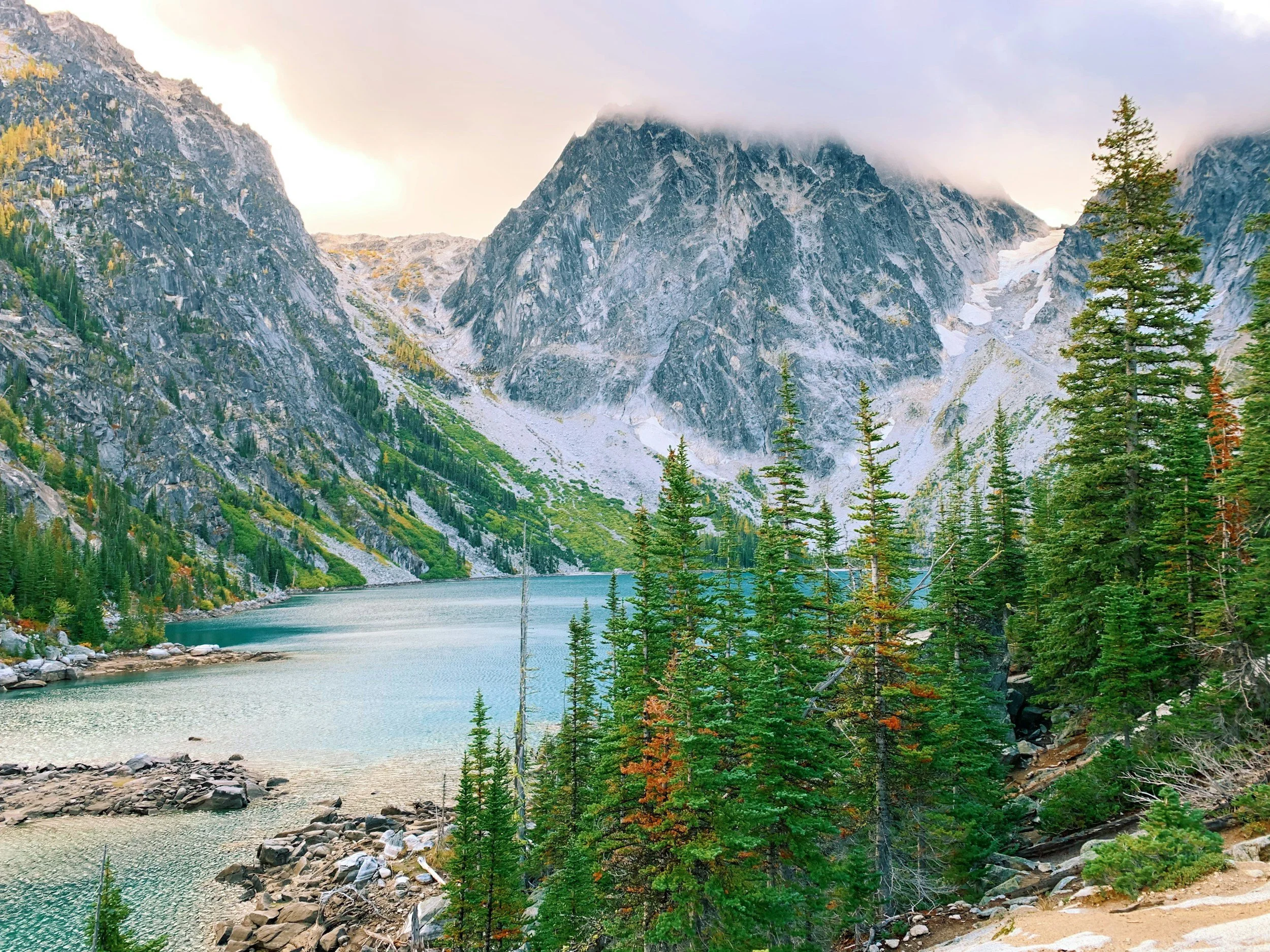 Mountainous landscape with a clear blue lake surrounded by evergreen trees and rocky peaks under a cloudy sky.