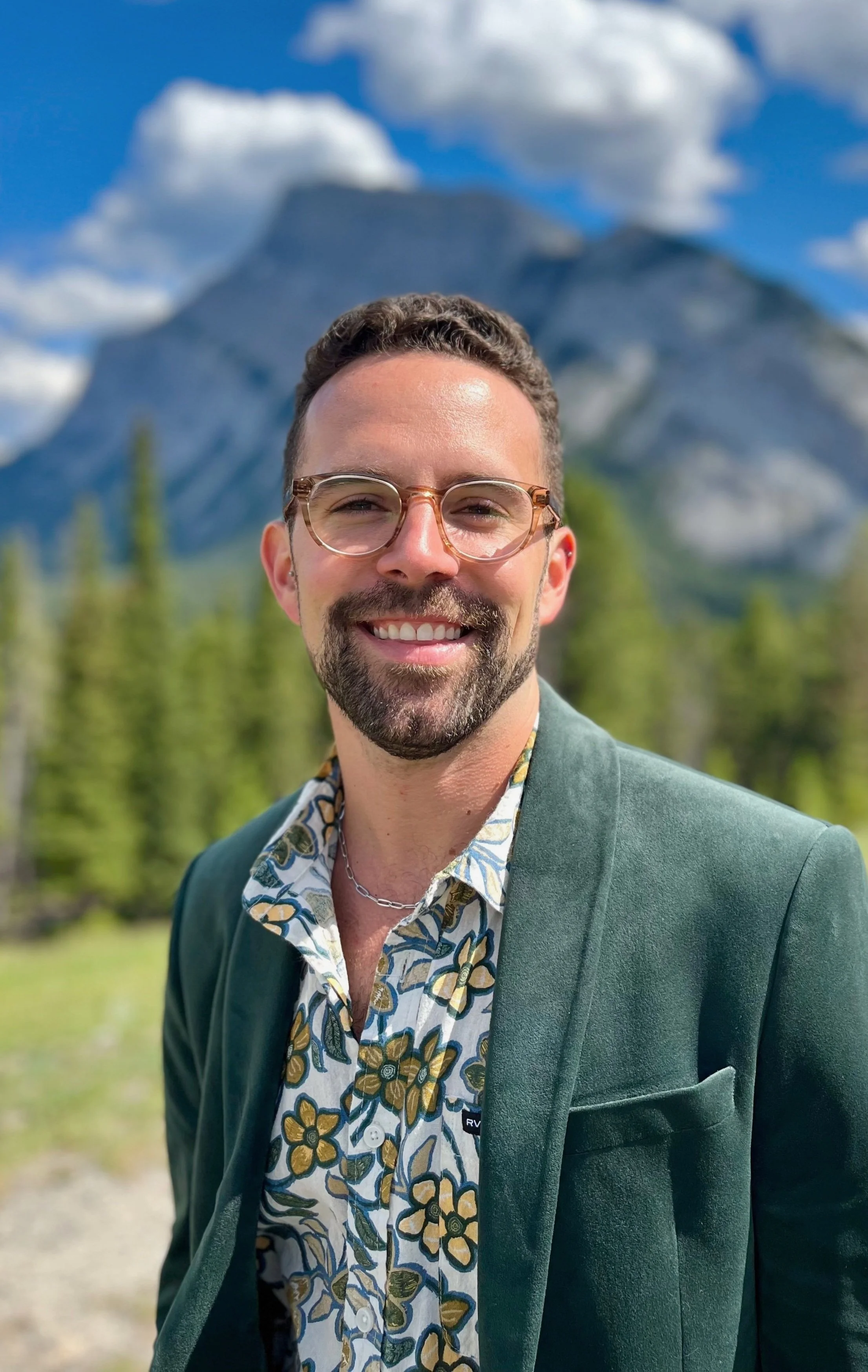 Smiling person with glasses, in a floral shirt and green jacket, standing outdoors in front of trees and mountains.
