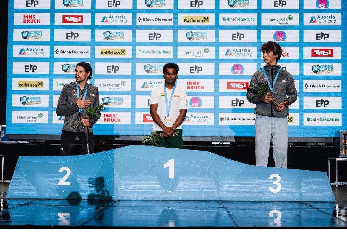 Three men standing on a podium at a climbing competition, Manikandan Kumar in 1st place, with sponsor logos in the background.