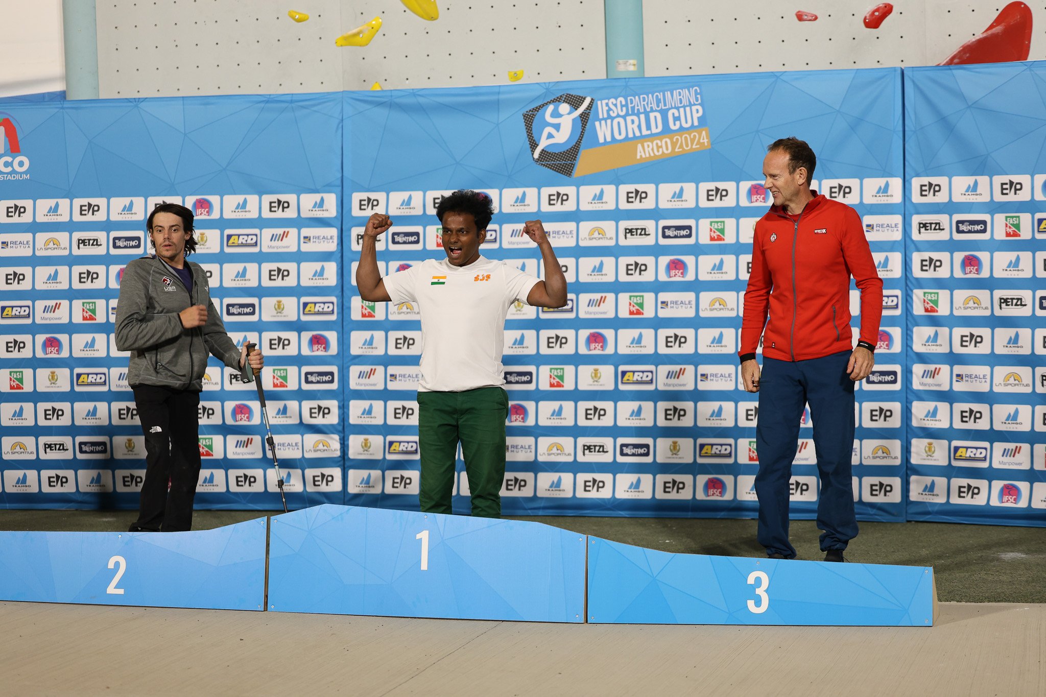 Three men on a podium at the IFSC Paraclimbing World Cup Arco 2024, with Manikandan Kumar in first place celebrating, second holding a walking aid, and third smiling.