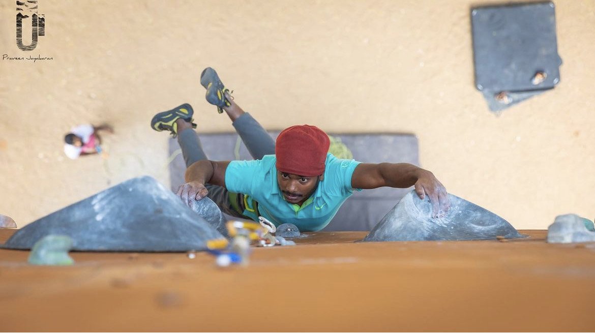 Manikandan Kumar climbing an indoor rock wall wearing a red cap and blue shirt, focusing intensely. Another climber is visible in the background.