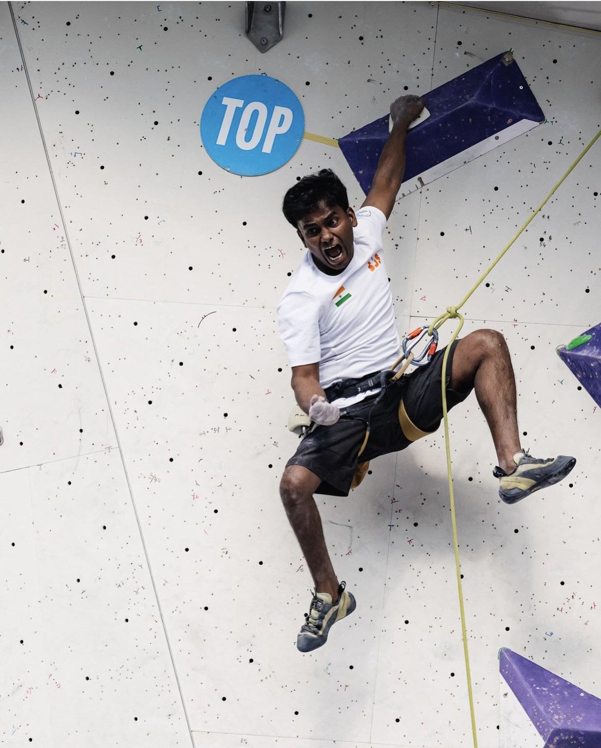 Manikandan Kumar reaching the top of an indoor climbing wall, excitedly celebrating with one hand holding a hold and the other in the air. The wall is marked with a 'TOP' sign.