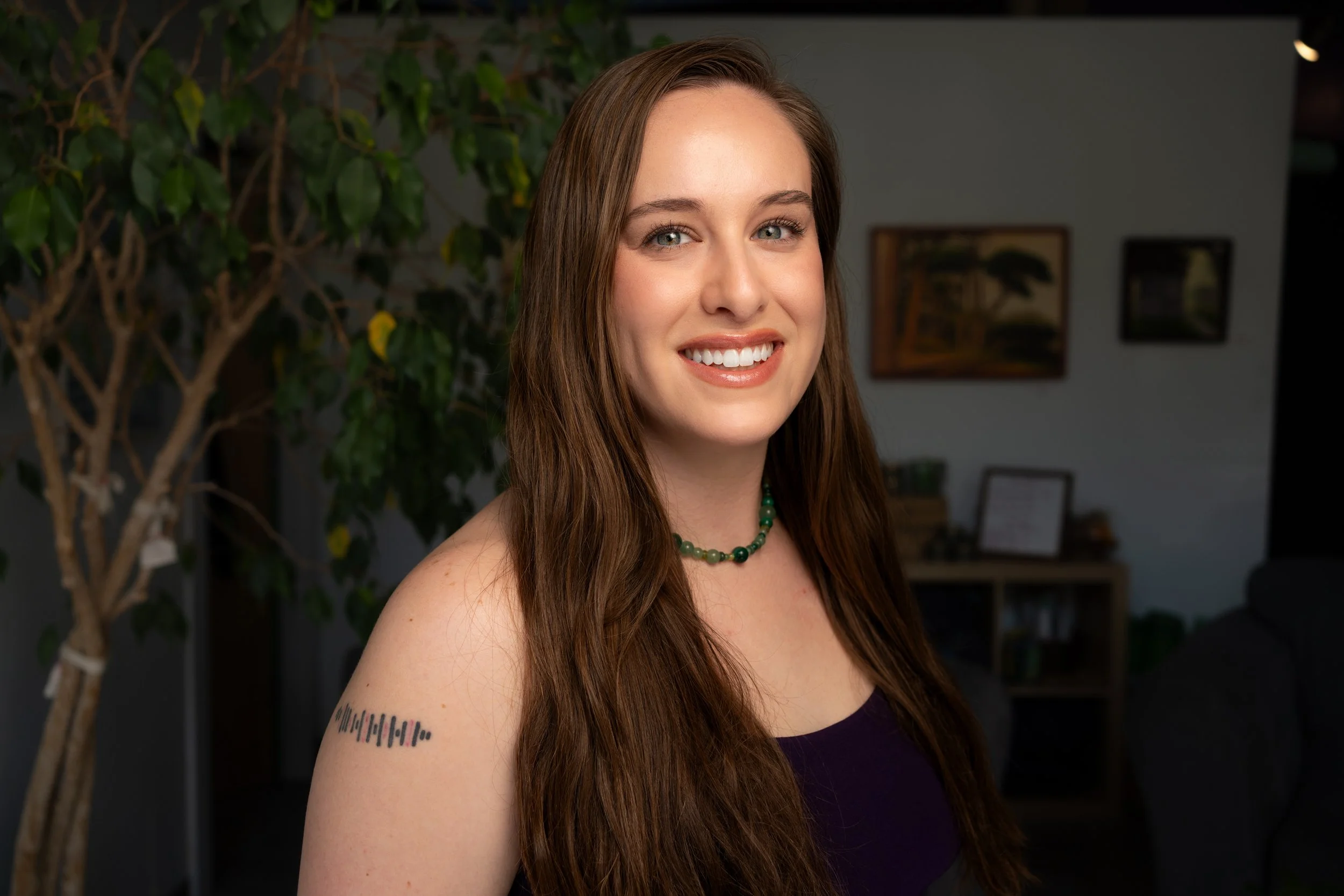 A woman with long brown hair, wearing a purple top and a green beaded necklace, smiling indoors with a background of a houseplant and framed artwork.