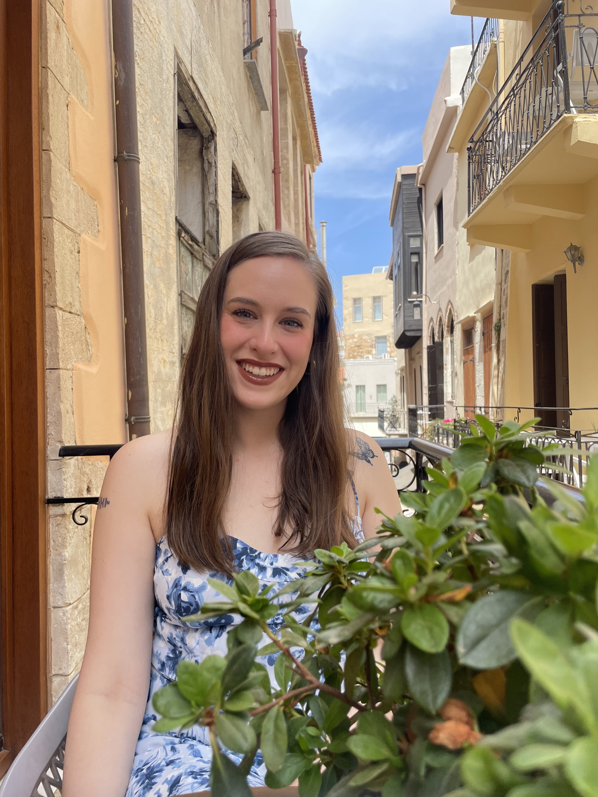 A woman with long brown hair and a floral dress smiling on a balcony in Greece with a view of colorful buildings and a blue sky.