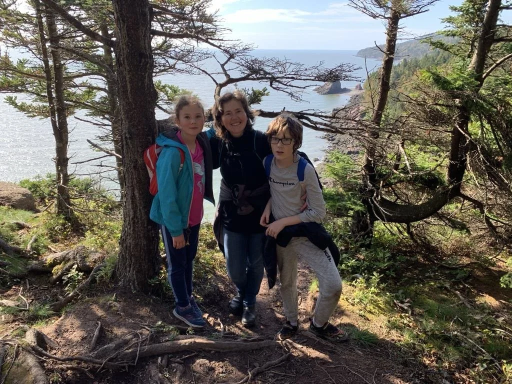 Armelle and her two children smiling while standing in a forest with a scenic ocean view in the background. One of her favorite spots: surrounded by nature, soaking in its calming and grounding benefits.